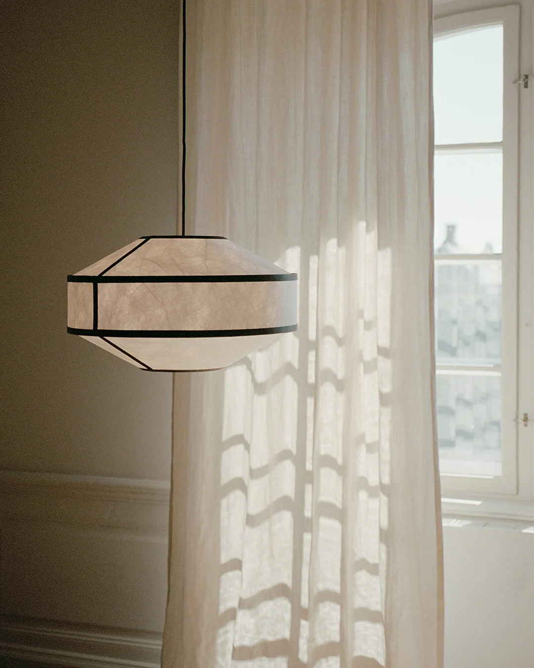 Eine geometrische Pendelleuchte aus Papier mit dunklen Rahmen hängt neben einem sonnenbeschienenen, durchsichtigen Vorhang und Fenster.