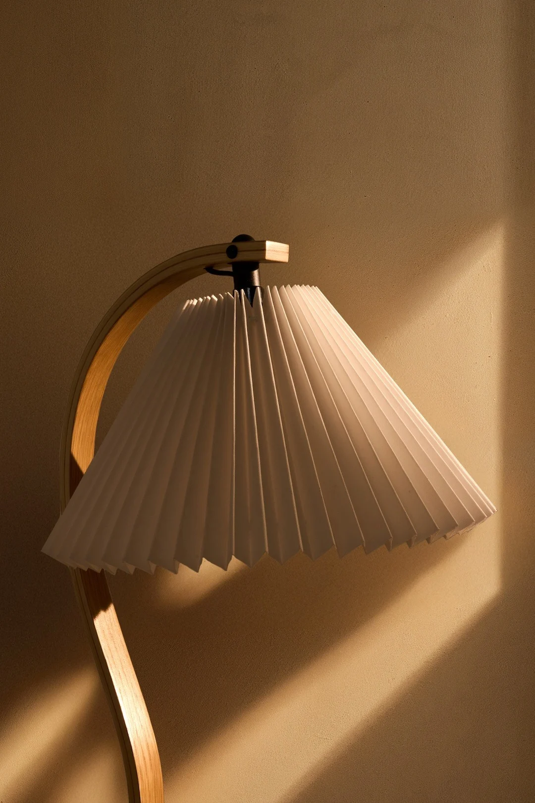 Eine Lampe mit einem weißen plissierten Schirm und einem gebogenen Holzfuß, die warmes Licht auf eine texturierte Wand wirft.