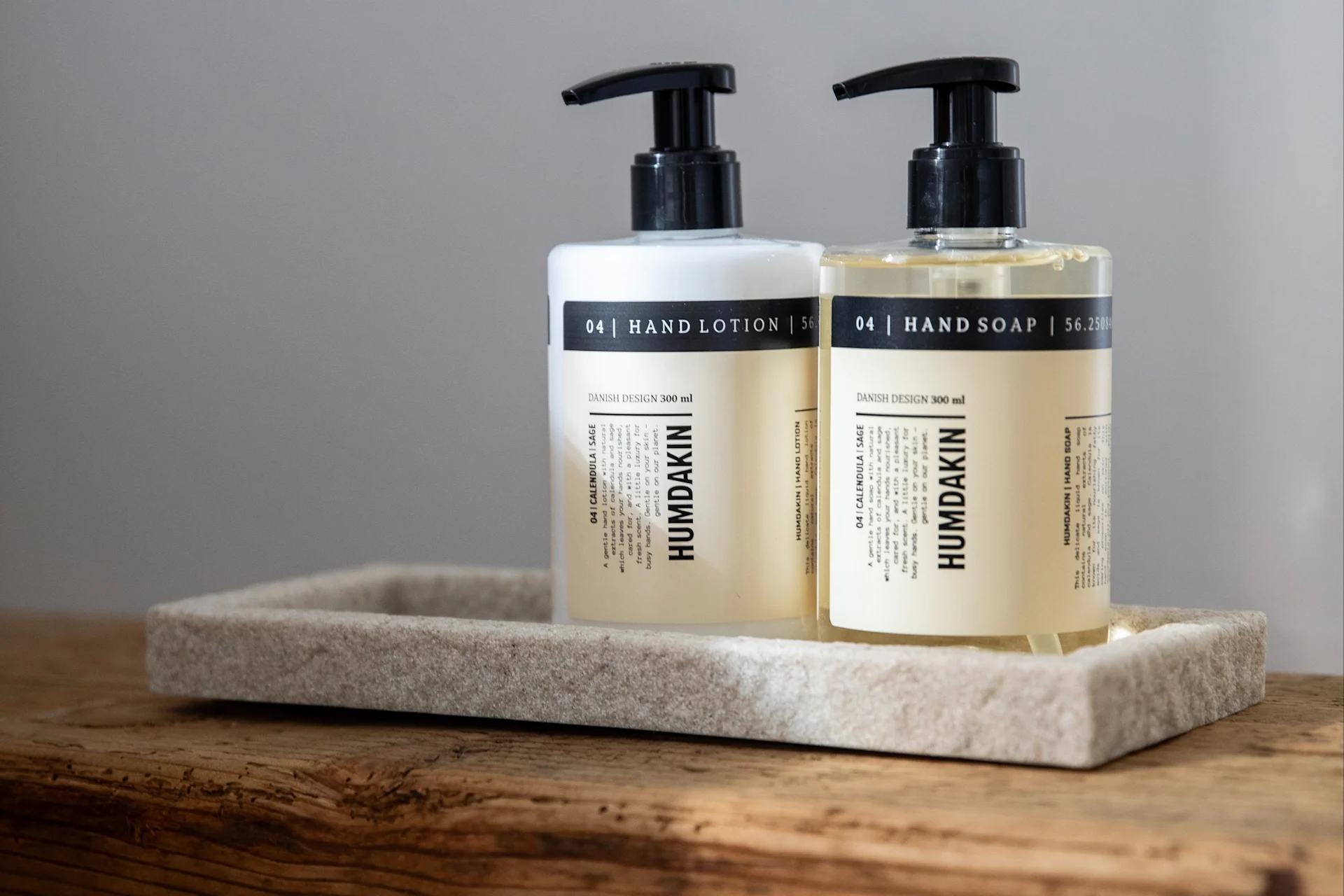 Zwei Humdakin Pumpflaschen, Handlotion (weiß) und Handseife (klar), auf einem steinähnlichen Tablett auf Holz.