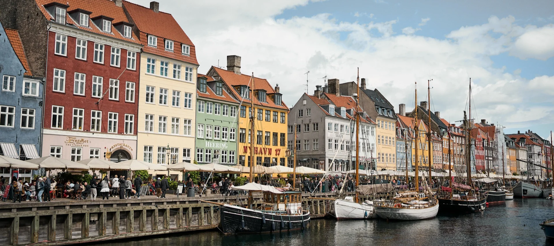 Bunte Gebäude an einem Kanal mit Booten und Menschen in Straßencafés unter bewölktem Himmel.