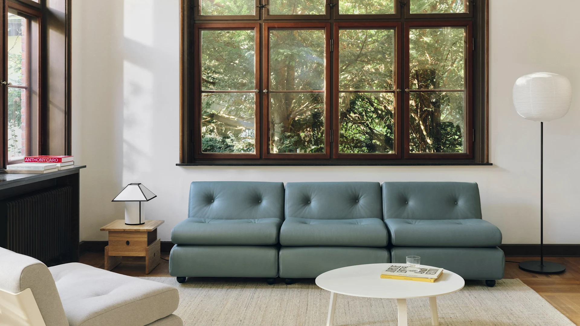 Ein modernes Wohnzimmer mit einem blau-grauen Modulsofa, weißem Couchtisch und einem großen Fenster mit Blick auf grüne Bäume. Zwei Lampen beleuchten den Raum.