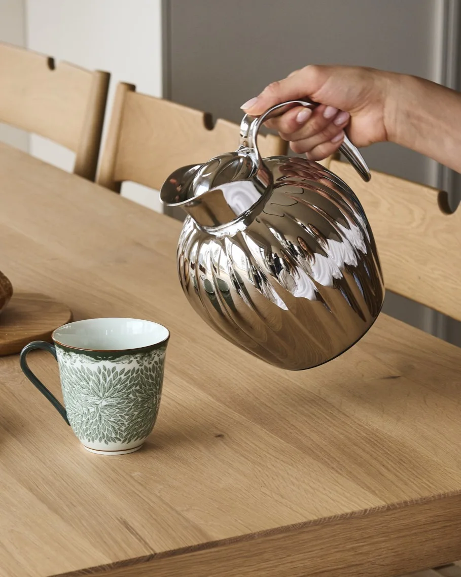 Eine Hand gießt aus einer silbernen geriffelten Karaffe in einen weißen Becher mit grünem Blattmuster auf einem Holztisch.