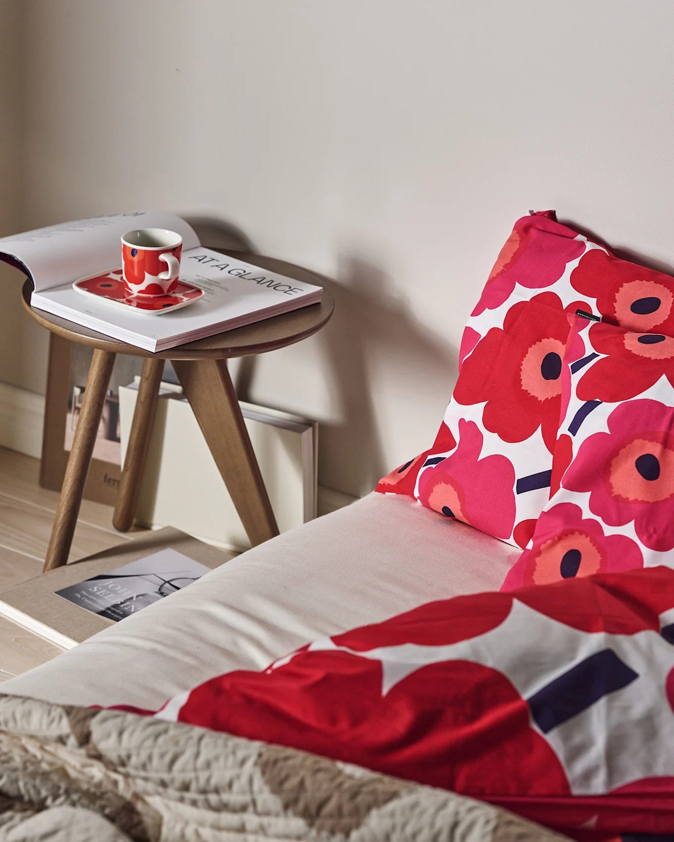 Ein Bett mit rot-rosa Blumenbettwäsche und einer strukturierten beigen Decke. Eine rote Tasse und ein offenes Buch auf einem Holzbeistelltisch.