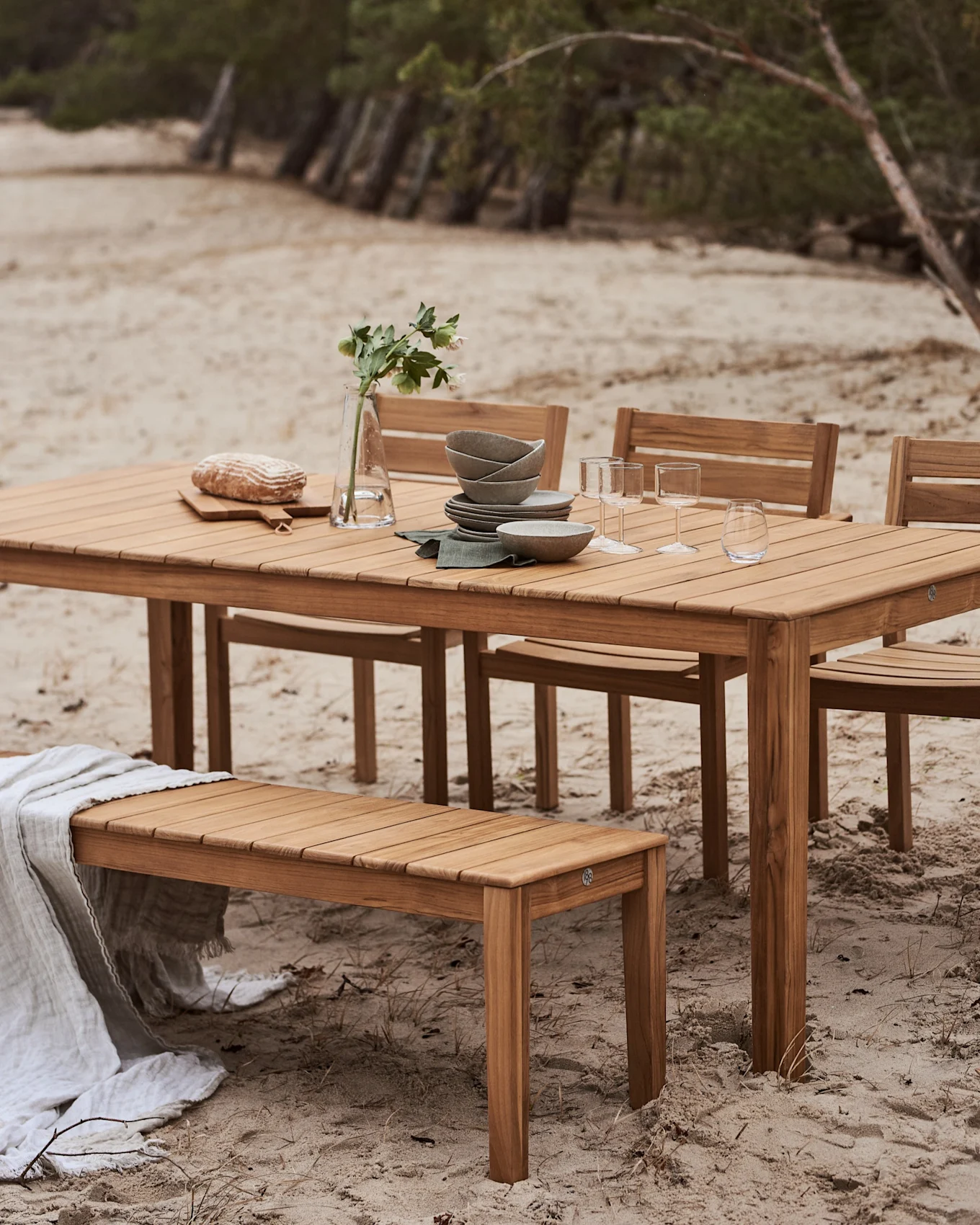 Outdoor-Essgruppe aus Holz mit Tisch und Bank an einem Sandstrand. Der Tisch ist mit Geschirr, Gläsern, Brot und einer Blumenvase gedeckt.