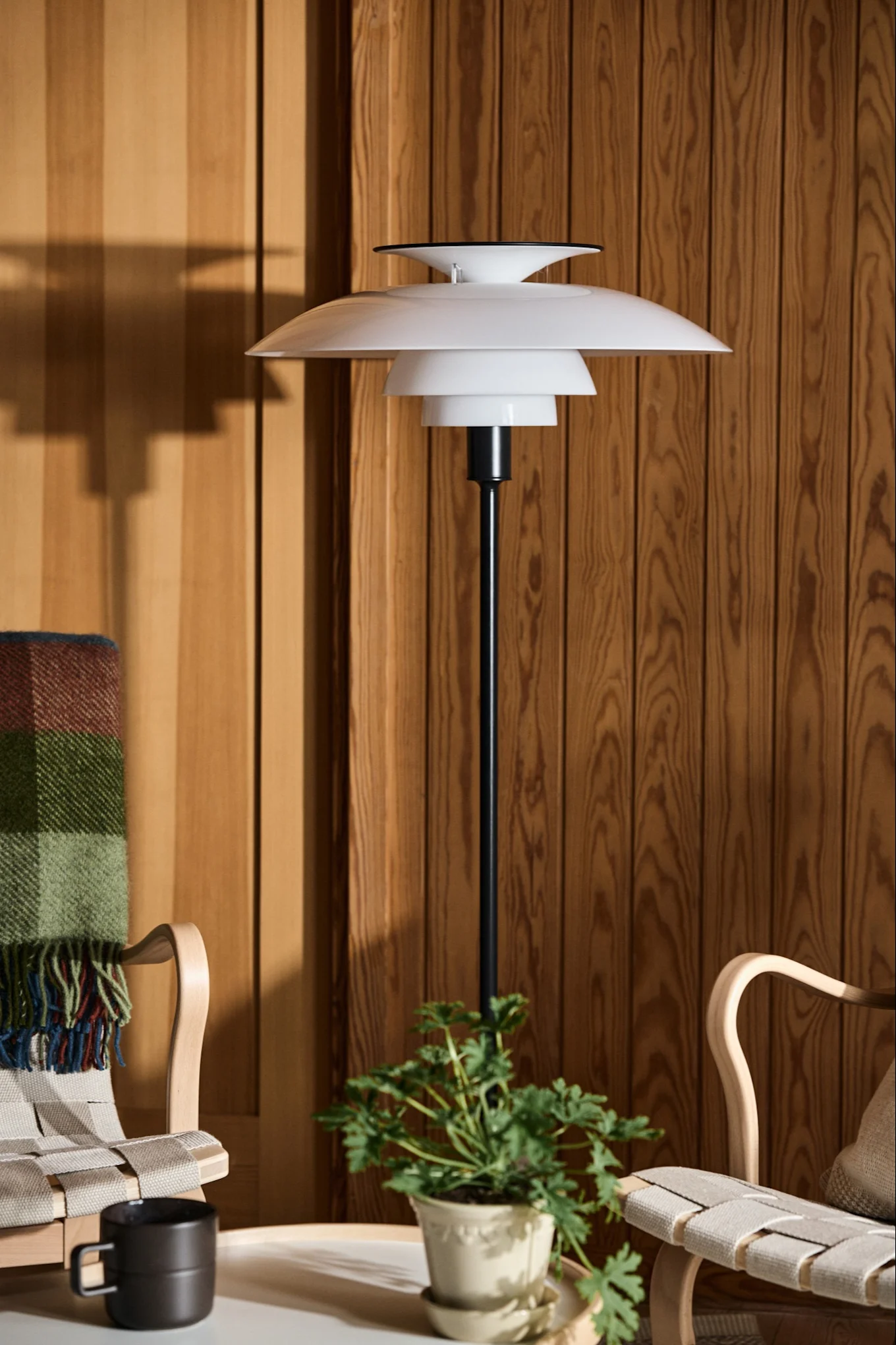 Weiße geschichtete Stehlampe neben zwei Holzstühlen und einer Pflanze vor einer holzvertäfelten Wand.