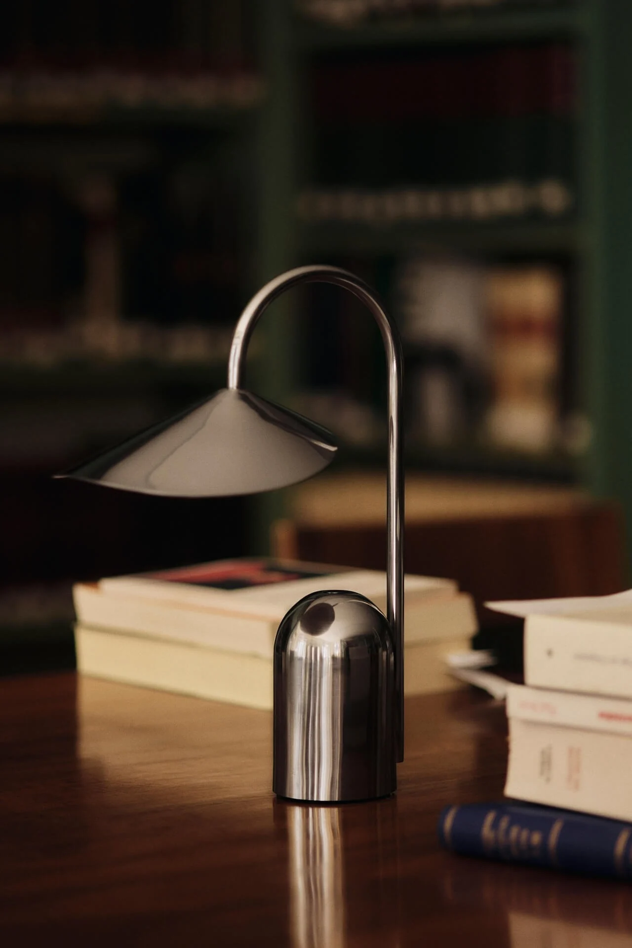 Eine moderne silberne Tischlampe mit gebogenem Stiel und konischem Schirm steht auf einem Holzschreibtisch mit unscharfen Büchern im Hintergrund.