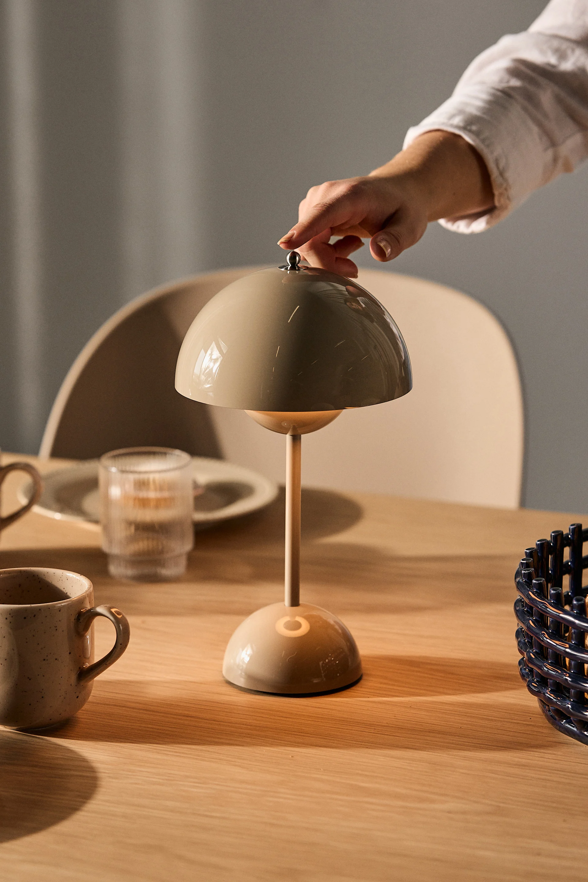Eine Hand stellt eine beige pilzförmige Tischlampe auf einem Holztisch mit Tassen und einem Glas ein.