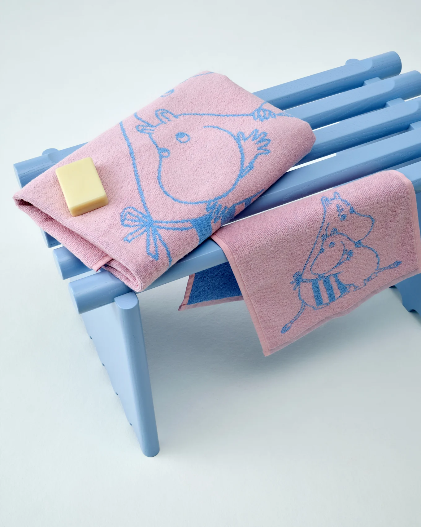 Zwei pinkfarbene Moomin-Handtücher mit blauen Mustern, eines gefaltet mit einem Stück Seife, auf einer hellblauen Bank.