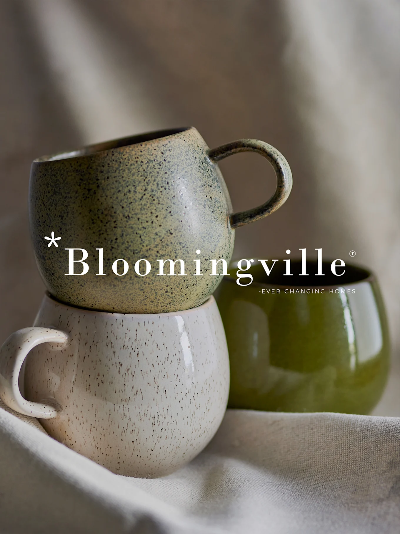 Bloomingville-Logo über einem Stapel von zwei gesprenkelten Keramikbechern in Grün und Cremeweiß, mit einem weiteren einfarbigen grünen Becher daneben.