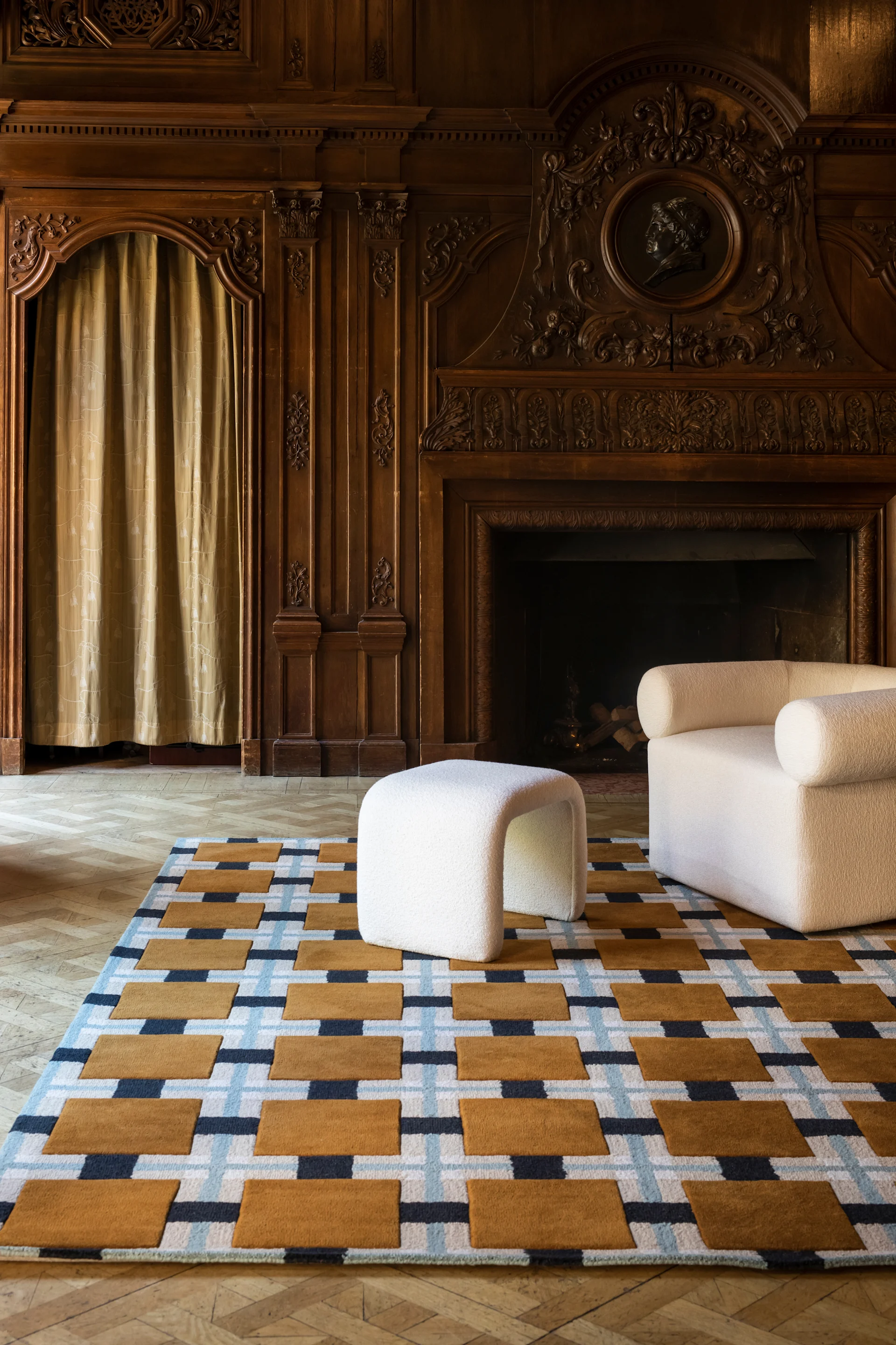 Geometrischer Teppich mit braunen, blauen, weißen Mustern, ein weißer strukturierter Hocker und Sessel in einem holzgetäfelten Raum.