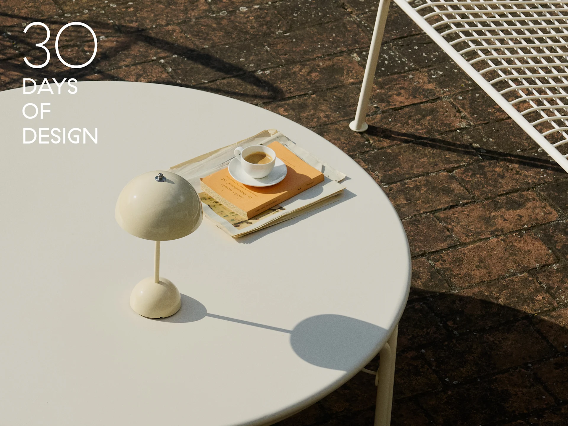 Cremefarbene Pilzlampe, Kaffeetasse und Buch auf einem weißen runden Gartentisch.