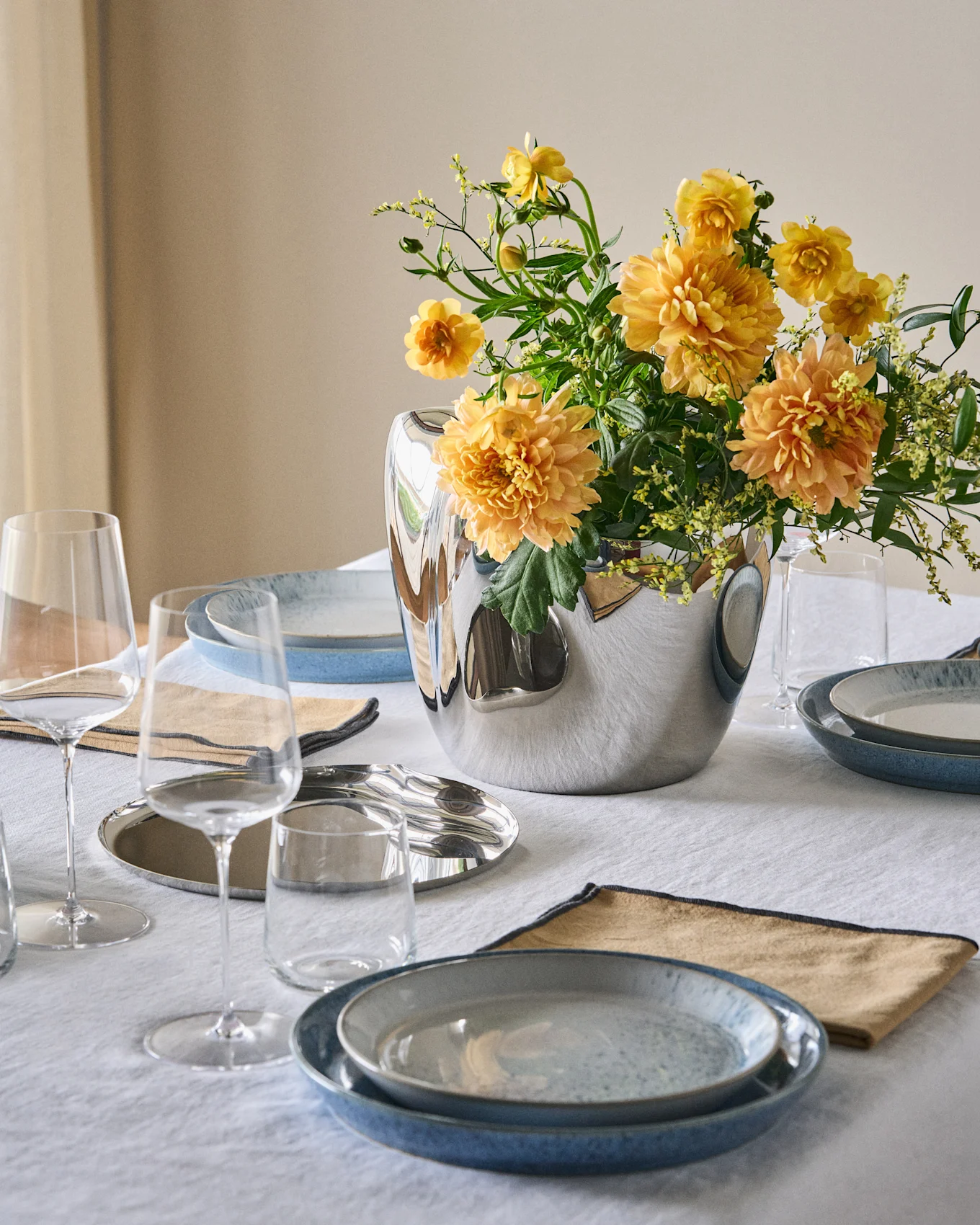 Ein gedeckter Esstisch mit blauen Tellern, beigen Servietten, klaren Gläsern und einer silbernen Vase mit orange-gelben Blumen auf einem weißen Tischtuch.