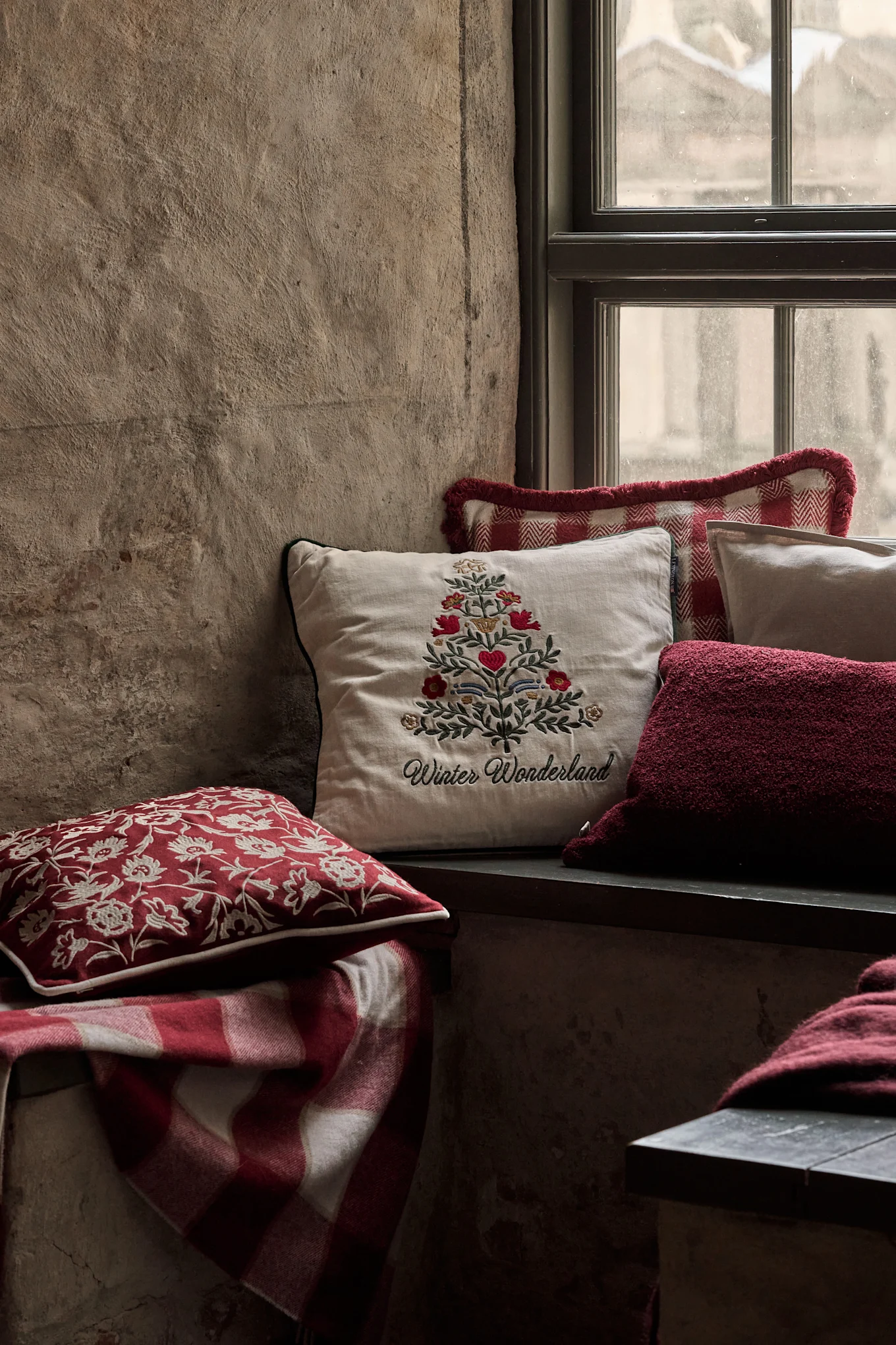 Gemütliche Fensternische mit festlichen Kissen, darunter eines mit einem bestickten Weihnachtsbaum und 'Winter Wonderland', und einer rot karierten Decke.