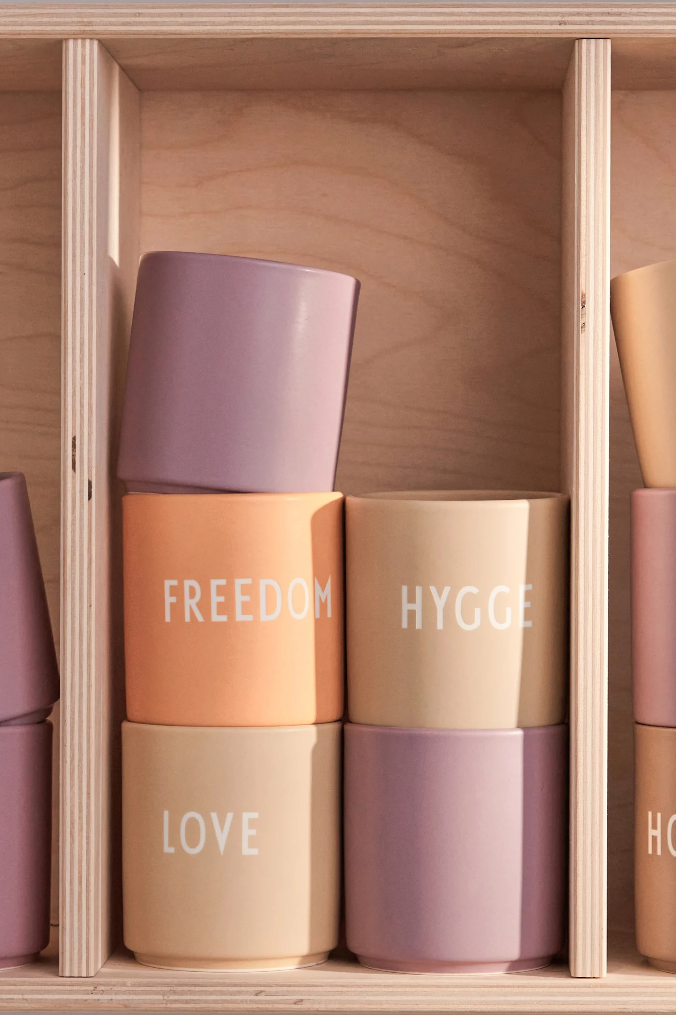 Gestapelte pastellfarbene Tassen in einem Holzregal. Tassen zeigen die Worte 'FREEDOM', 'HYGGE' und 'LOVE' in weißer Schrift.