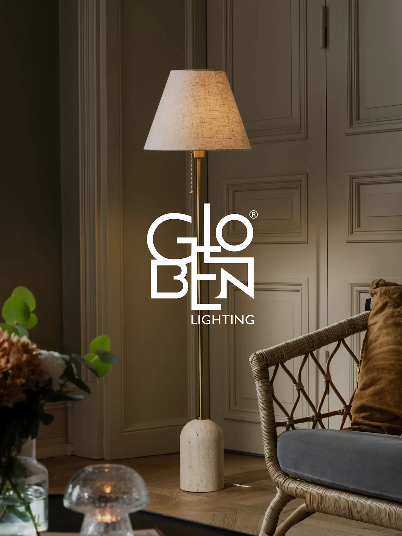 Eine Stehlampe mit beigem Schirm, Messingständer und abgerundetem Steinsockel steht neben einem Korbstuhl.
