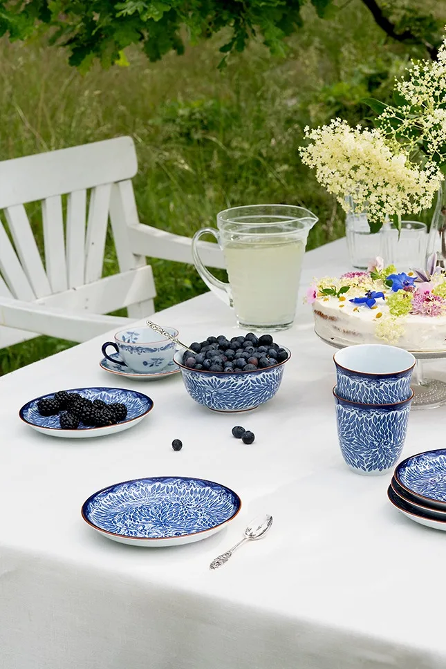 Gartentisch mit blau-weißem Geschirr, frischen Heidelbeeren und Brombeeren, Limonadenkrug und blumengeschmückter Kuchen.
