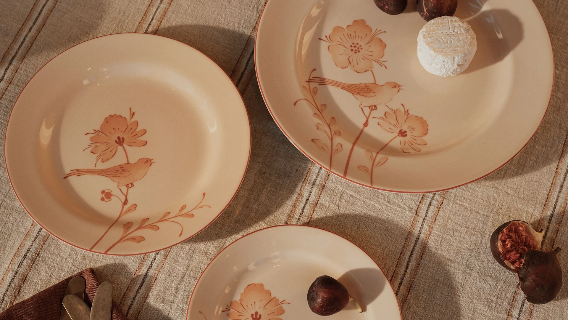 Drei hellpfirsichfarbene Teller mit Vogel- und Blumenmotiven auf einem gestreiften Tischtuch, mit Feigen und einem Käse.