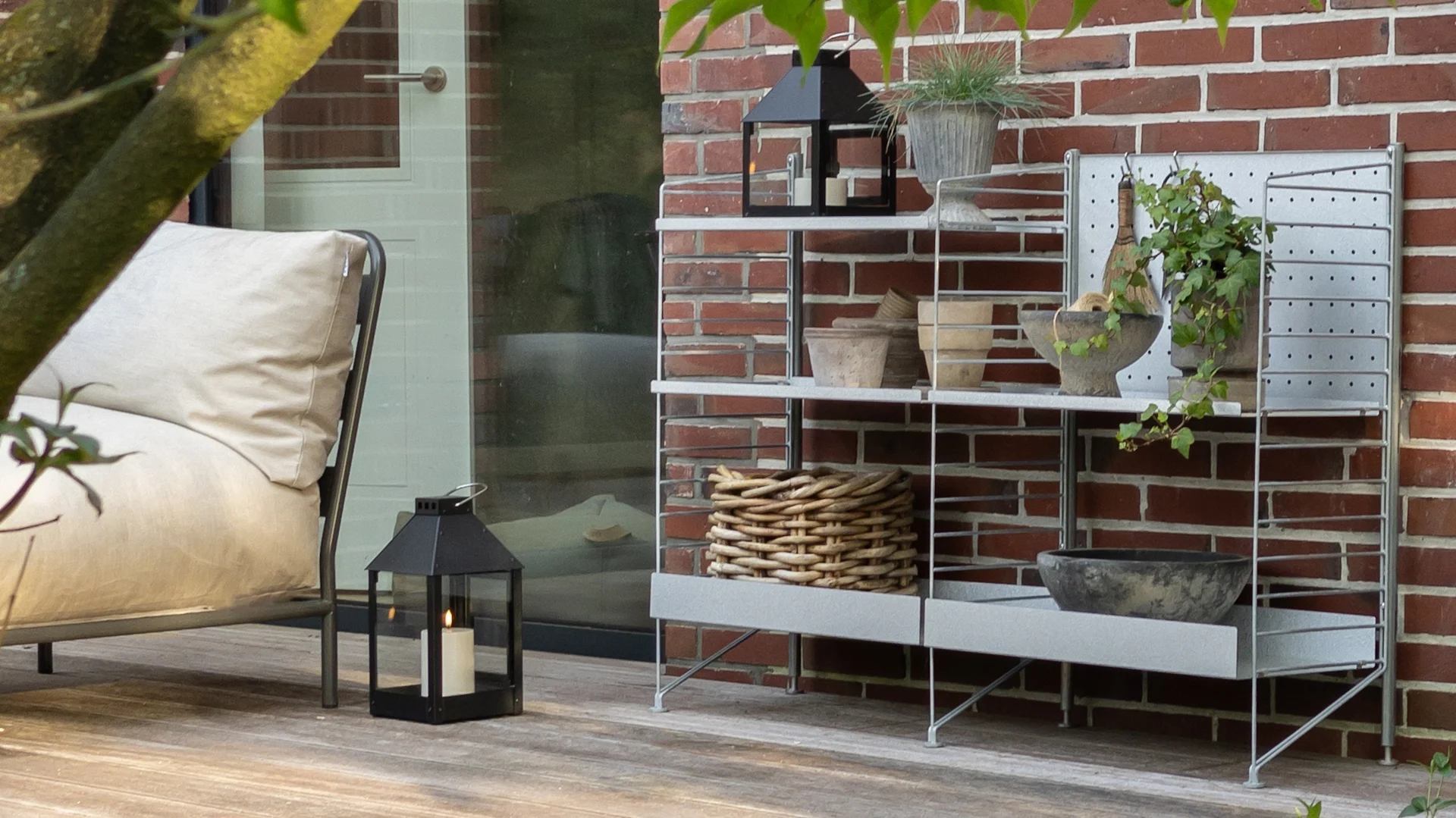 Eine Außenterrassenszene mit einem hellgrauen Regal vor einer roten Ziegelwand, das Pflanzen, Töpfe, einen geflochtenen Korb und Laternen enthält. Ein Loungesessel und eine Laterne stehen auf der Holzterrasse.