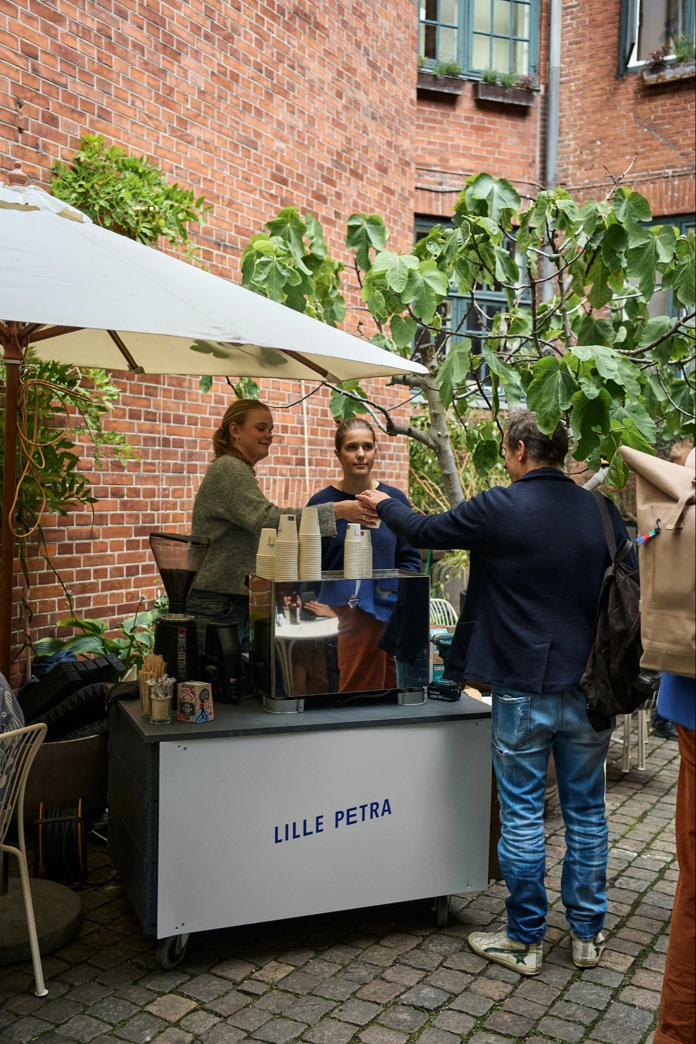 Zwei Frauen an einem Outdoor-Kaffeestand bedienen einen Mann, vor Ziegelwänden und grünen Pflanzen.