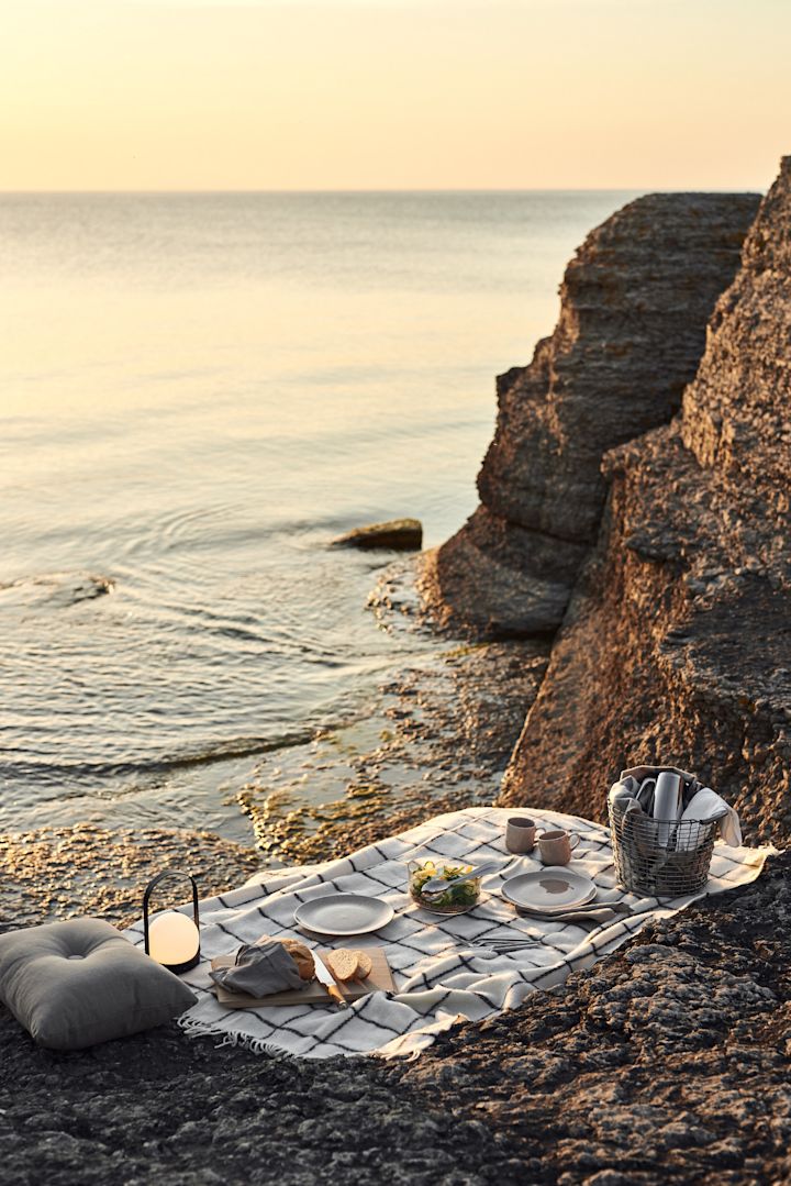 Summer Bucket List: Eine Picknickdecke liegt am Strand, darauf befinden sich Teller, Essen, eine tragbare Leuchte und ein Korb.