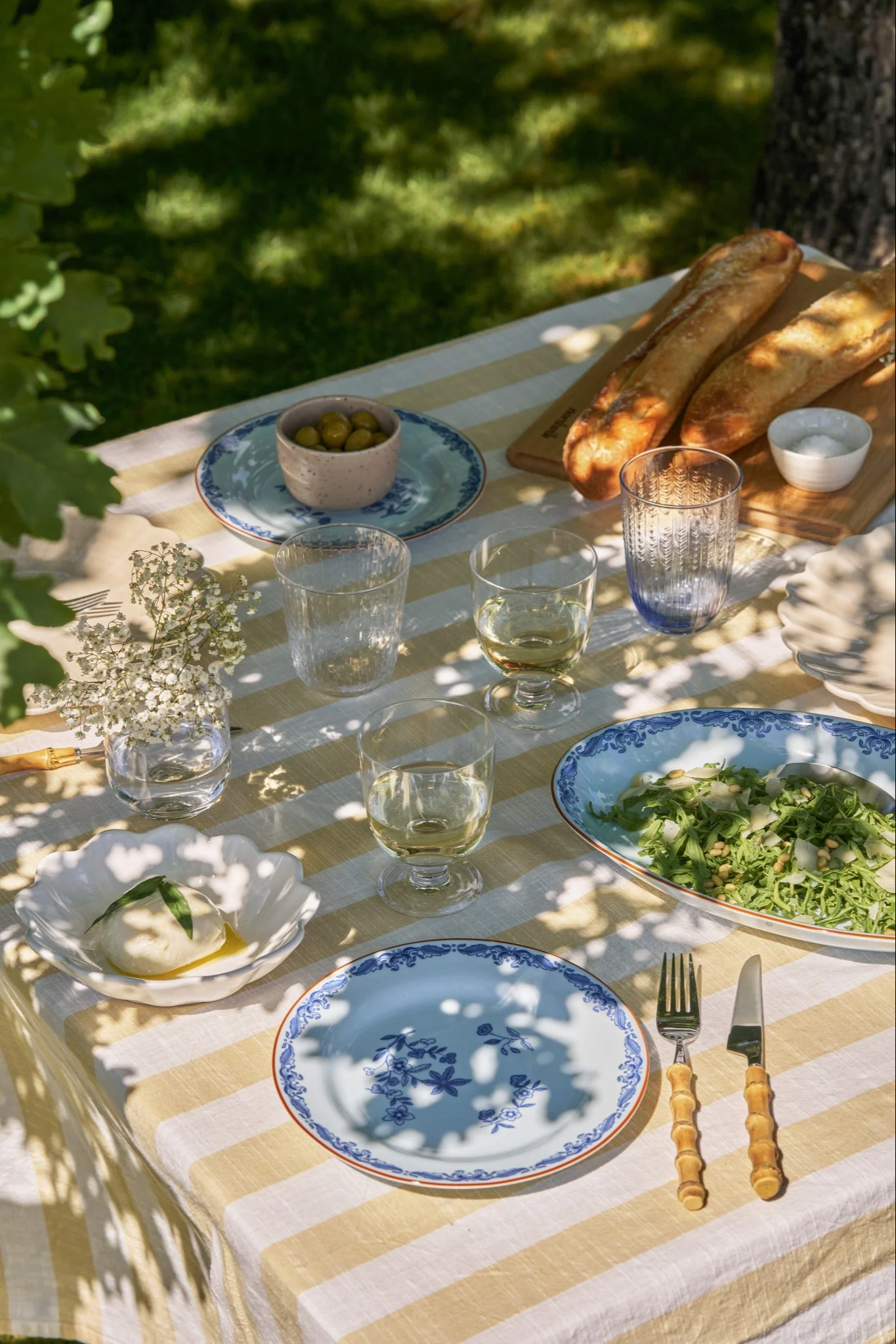 Ein Outdoor-Picknicktisch mit gelb-weiß gestreifter Tischdecke, Burrata, Salat, Baguettes, Oliven und blau gemusterten Tellern.
