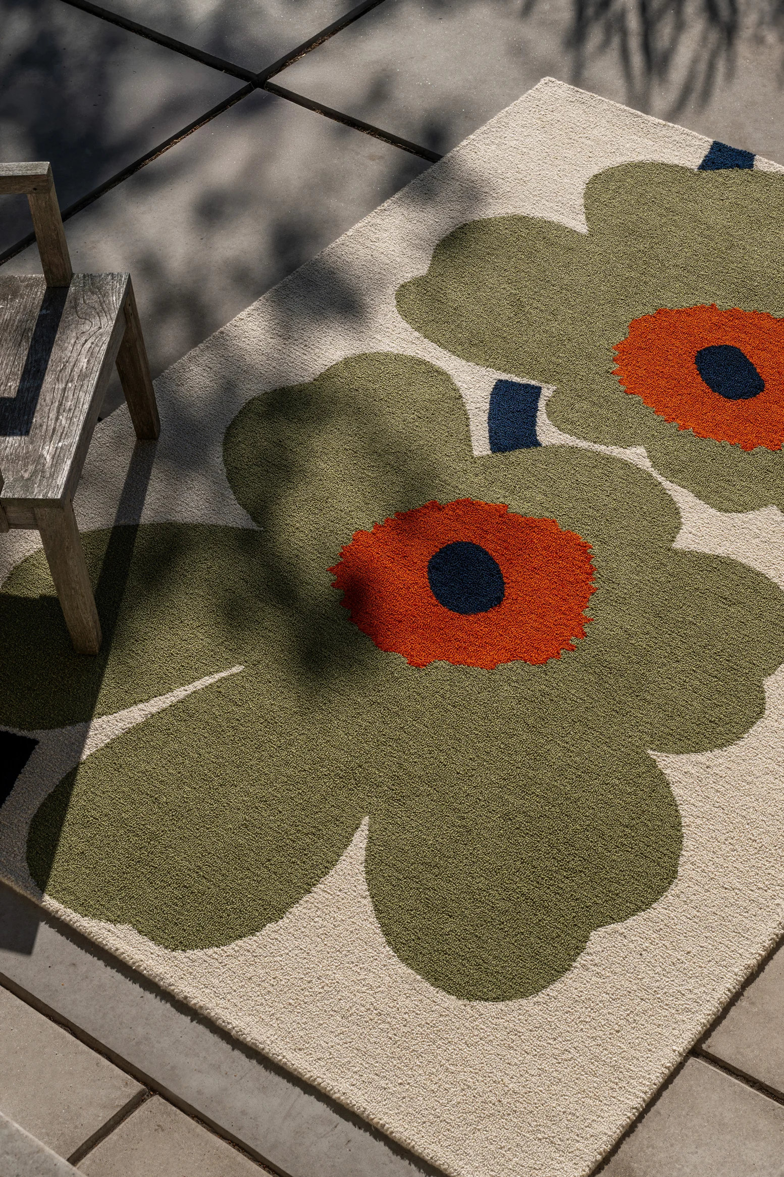 Teppich mit großen grünen und orangefarbenen Blumen auf hellem Hintergrund, unter Holzstuhl auf Terrasse, mit Blattschatten.