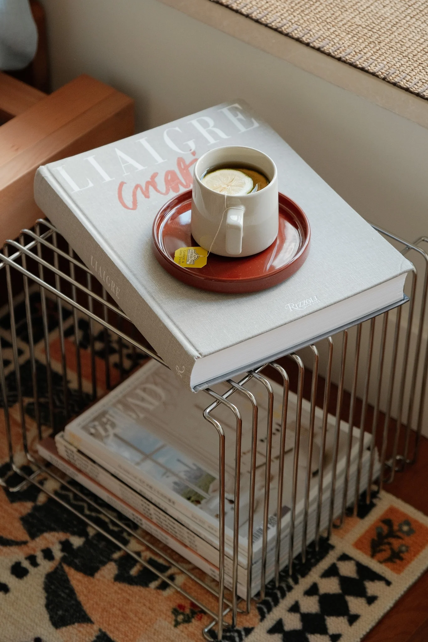 Eine weiße Tasse mit Zitronentee auf einem roten Untersetzer liegt auf einem hellgrauen Buch auf einem Drahtregal.