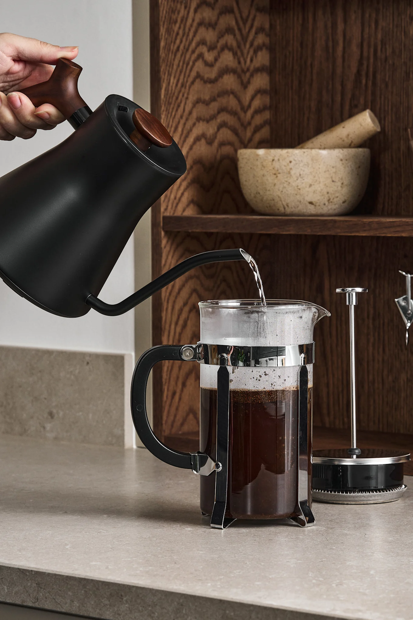 Eine Hand gießt heißes Wasser aus einem schwarzen Schwanenhalskessel in eine French Press mit Kaffeepulver auf einer Arbeitsplatte.