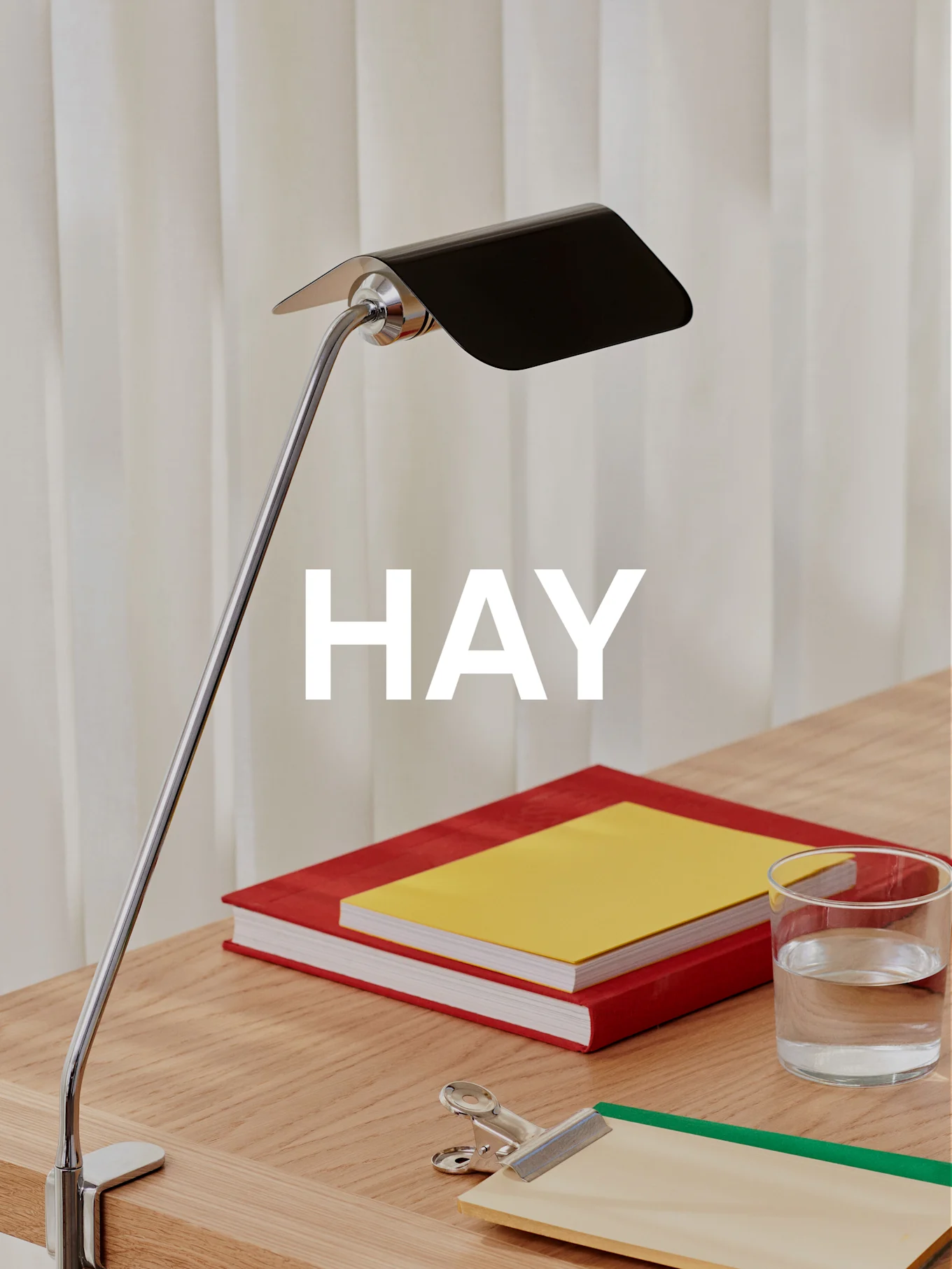 Eine Chrom-Schreibtischlampe mit schwarzem Schirm beleuchtet einen Holzschreibtisch mit zwei Büchern, einem Glas Wasser und einem Klemmbrett.
