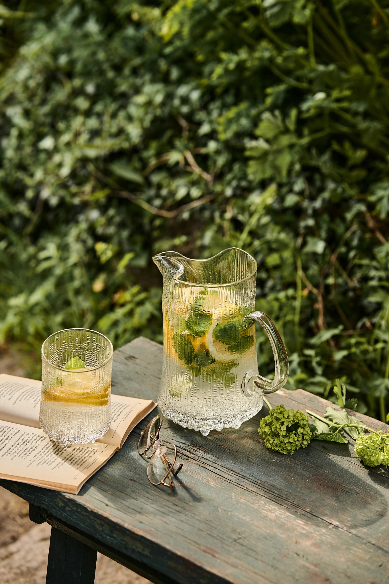 Der Ultima Thule Krug und das passende Glas von Iittala stehen auf einer Bank im Sonnenschein mit einem Buch. Holunderblüten-Sirup selbst zu machen, ist ein Muss für jede Summer Bucket List.