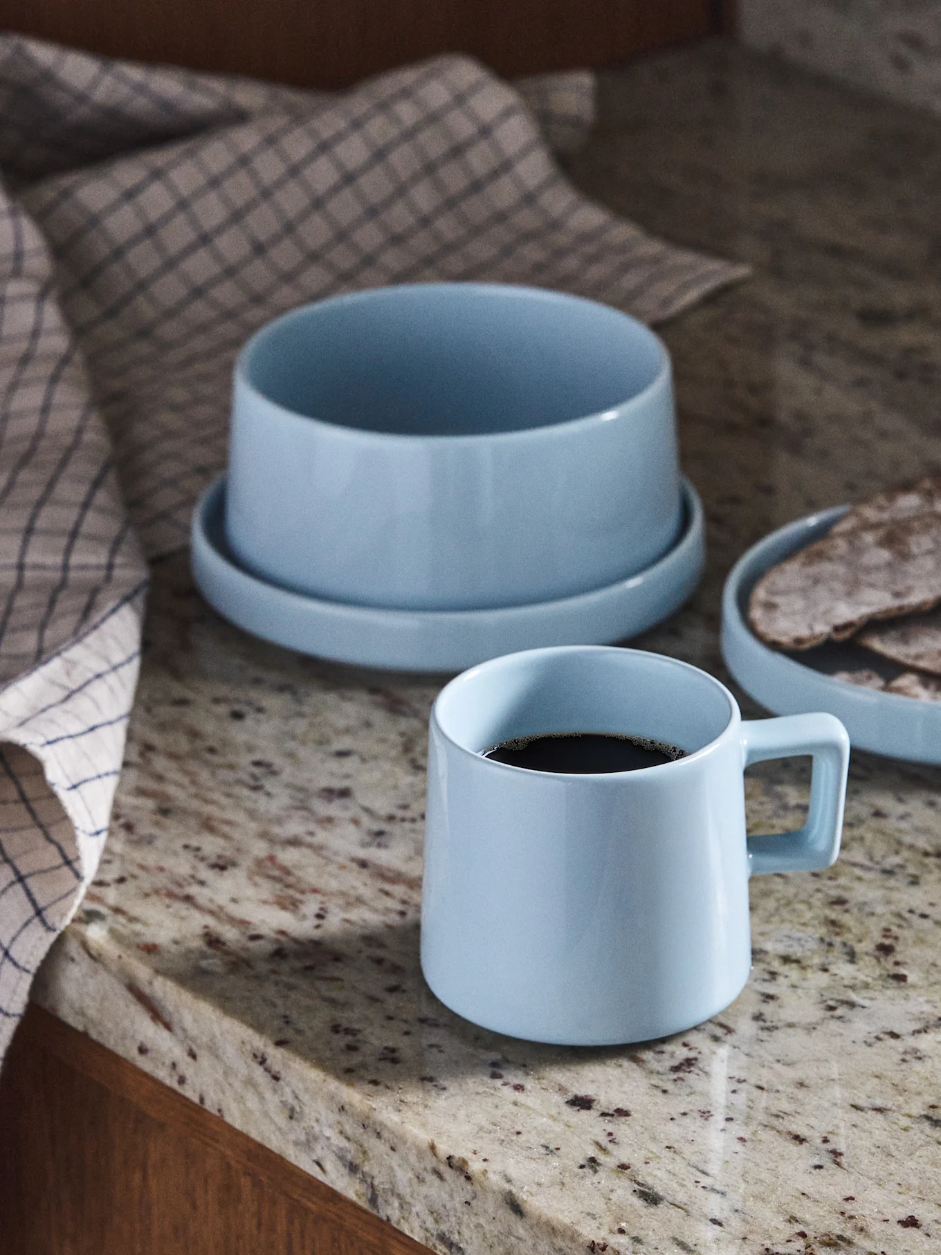 Hellblaue Keramiktasse mit Kaffee, Schale und Teller mit Fladenbrot auf einer Granitarbeitsplatte.