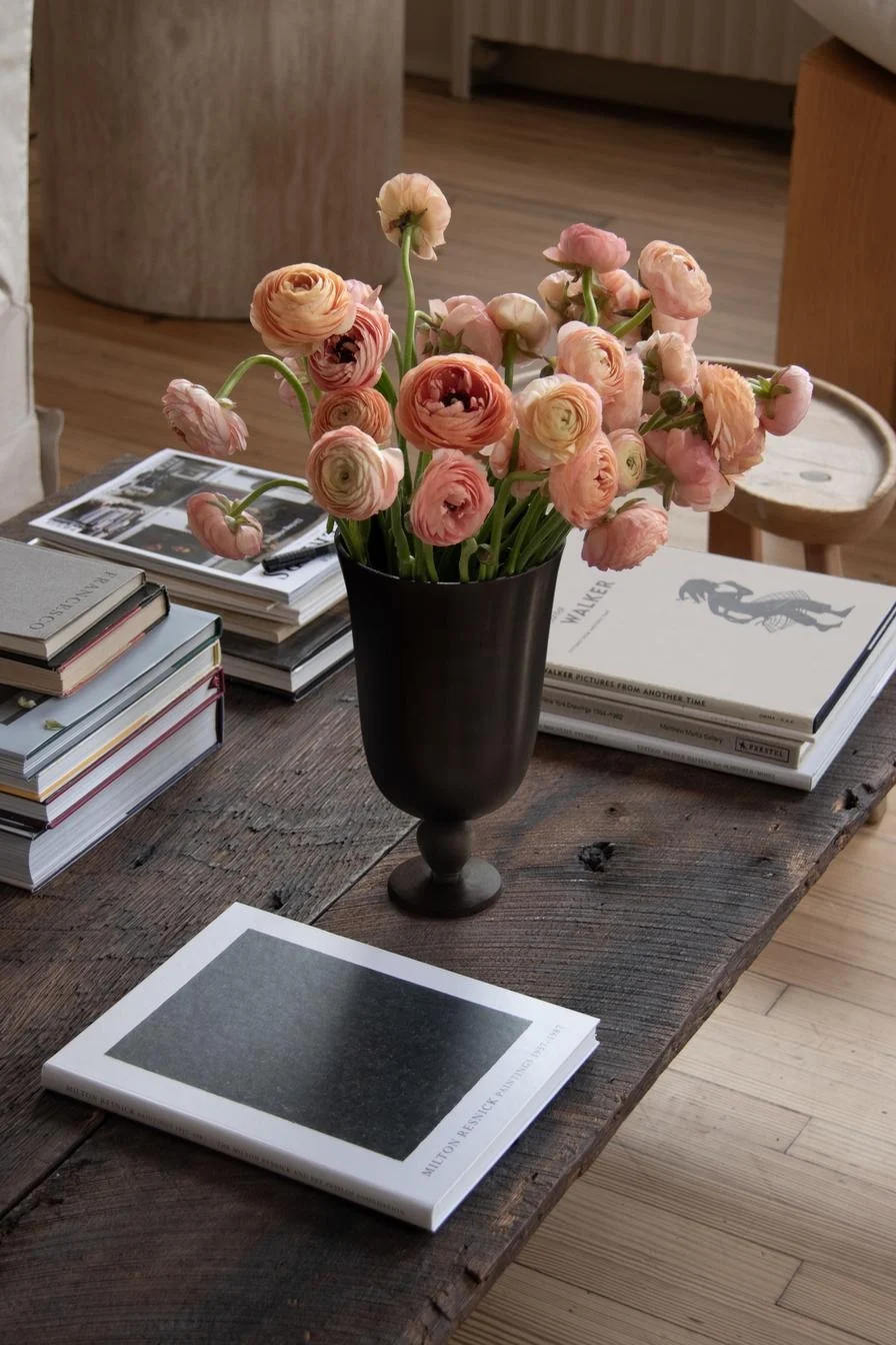 Pfirsichfarbene Ranunkeln in dunkler Vase auf Holztisch, umgeben von Kunstbüchern.