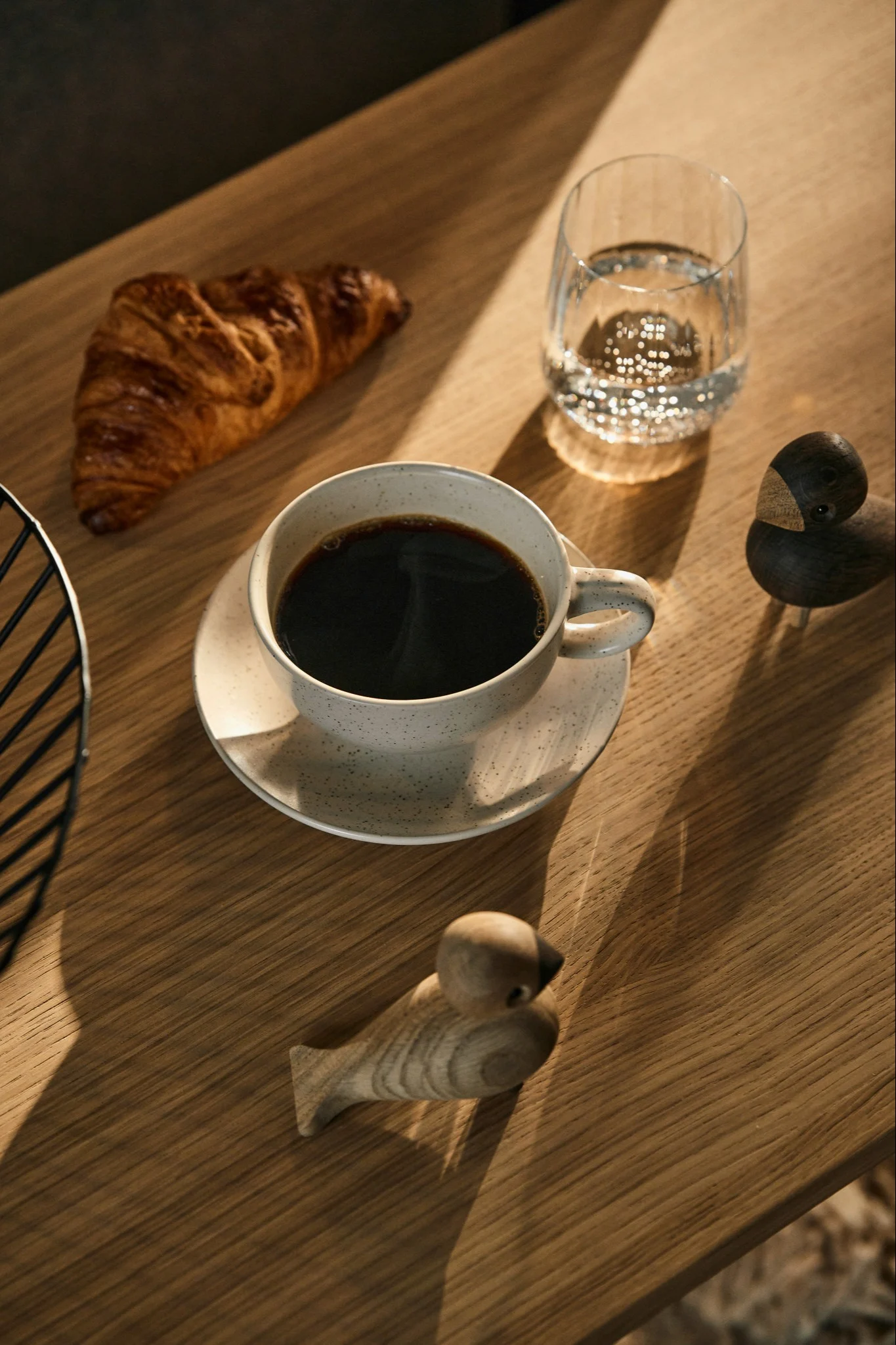 Ein Frühstückstisch mit Kaffee, einem Croissant, Wasser und zwei Holzfiguren auf einem sonnigen Holztisch.