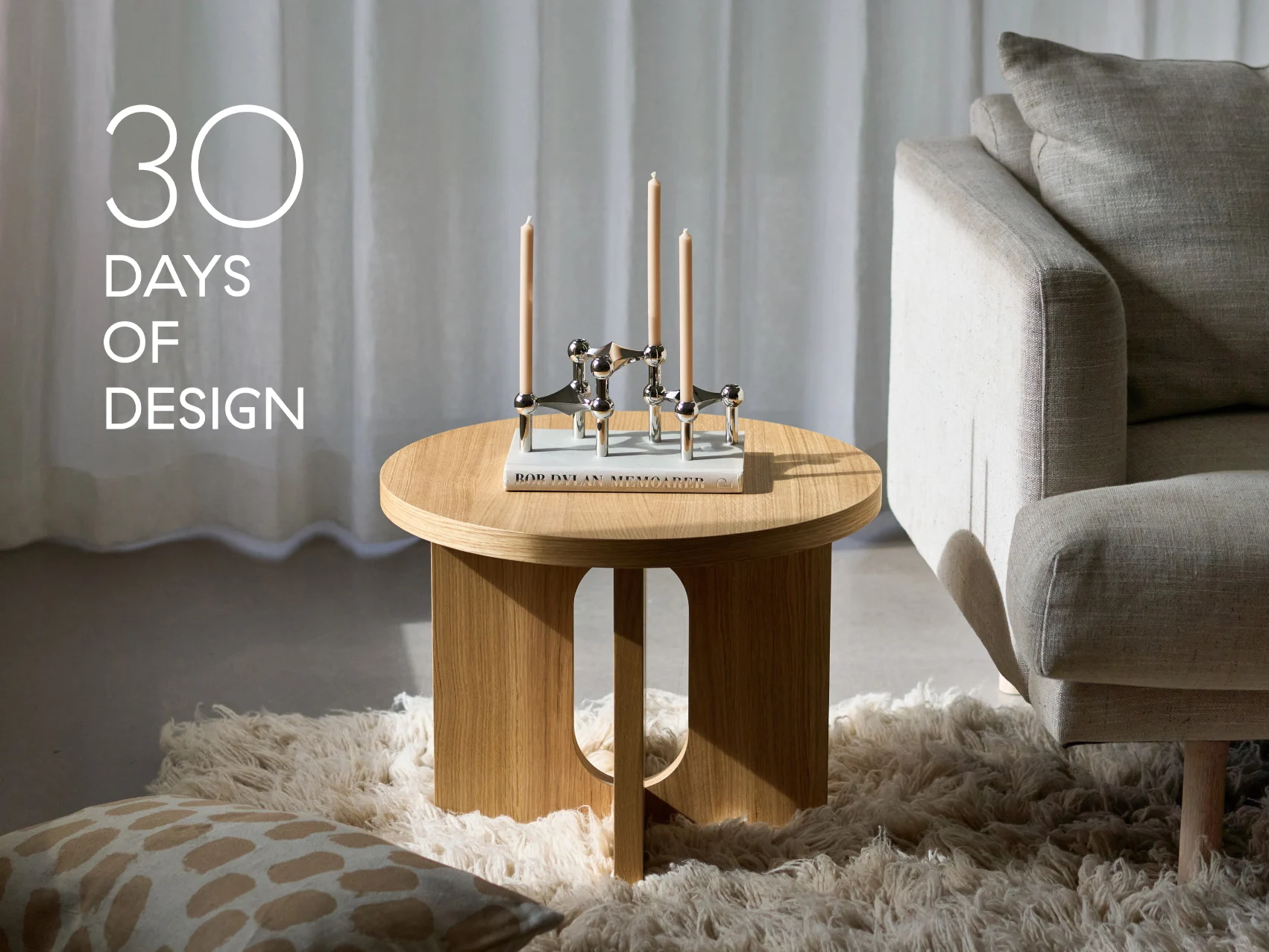 Modernes Wohnzimmer mit rundem Holztisch, silbernem Kerzenständer mit drei Kerzen, einem Buch und einem beigen Sofa. Text: '30 TAGE DESIGN'.