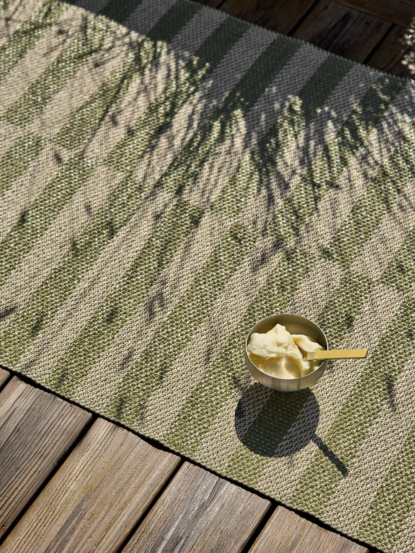 Eine silberne Dessertschale mit gelbem Löffel steht auf einer grün-beige gestreiften Outdoormatte auf einer Holzterrasse, gesprenkelt von Palmenschatten.