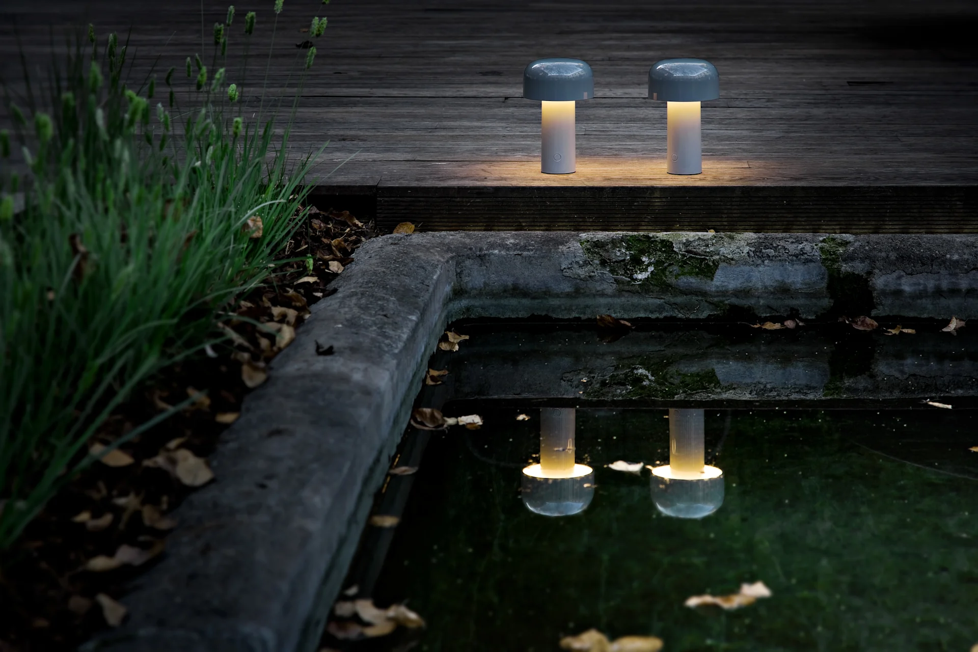 Zwei tragbare, pilzförmige Tischlampen mit warmem gelbem Licht stehen auf einem dunklen Holzdeck an einem Teich und spiegeln ihren Schein wider.