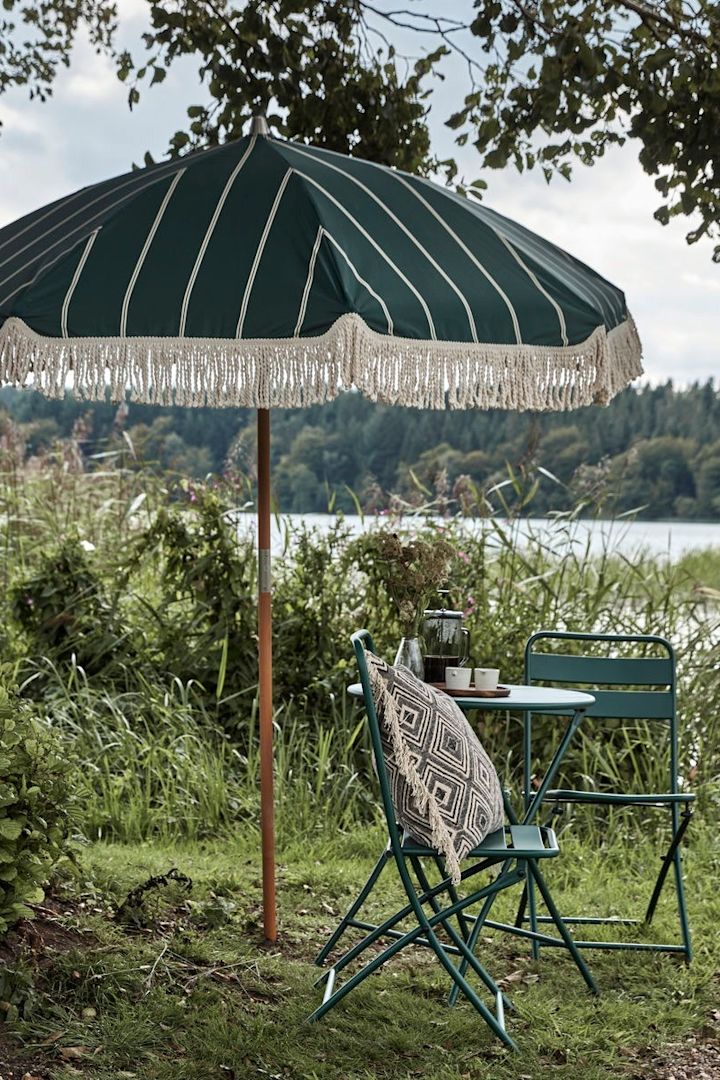 Boho Deko für den Garten: Der grüne Block Sonnenschirm mit Fransen von House Doctor in einem Garten, daneben eine kleine Sitzgruppe.