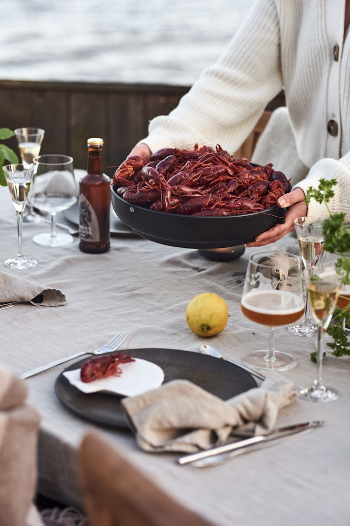 Wenn Sie noch nie eine schwedische Flusskrebs-Party erlebt haben, sollten Sie sie auf Ihre Summer Bucket List setzen. Hier sehen Sie ein Paar Hände, die eine Schale mit Flusskrebsen auf den Tisch stellen.