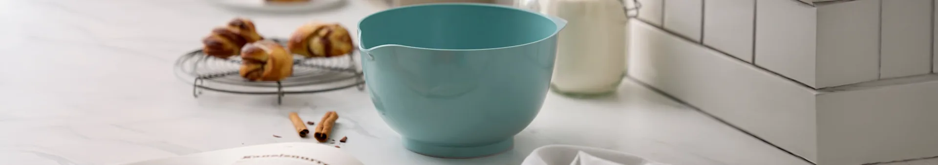 Hellblaue Rührschüssel, Gebäck auf Gitter, Milchflasche und Rezeptbuch auf Marmorarbeitsplatte.