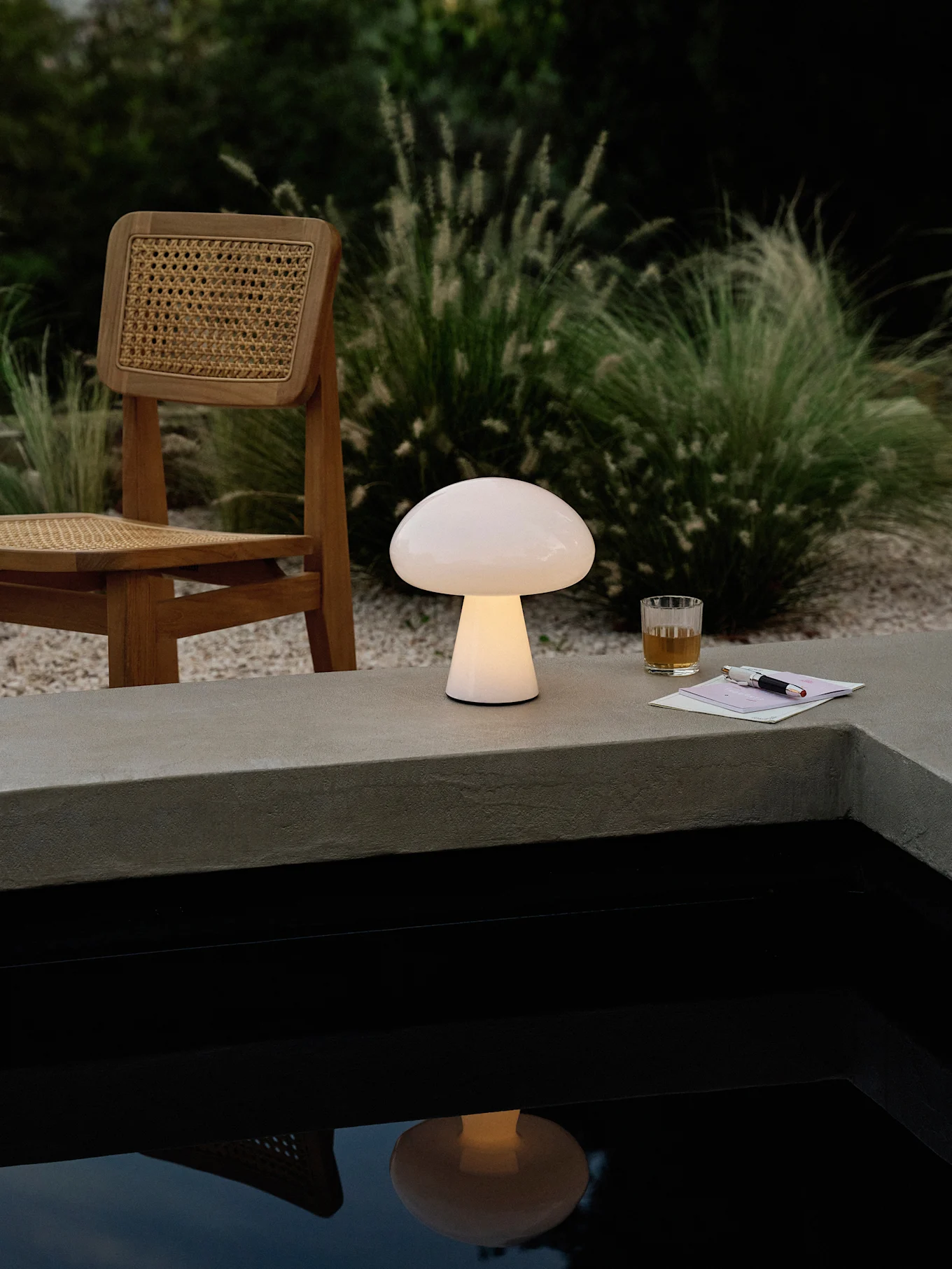 Eine leuchtende pilzförmige Lampe auf einem Betonsims neben einem Pool, mit einem Holzstuhl, Getränk und Stift.