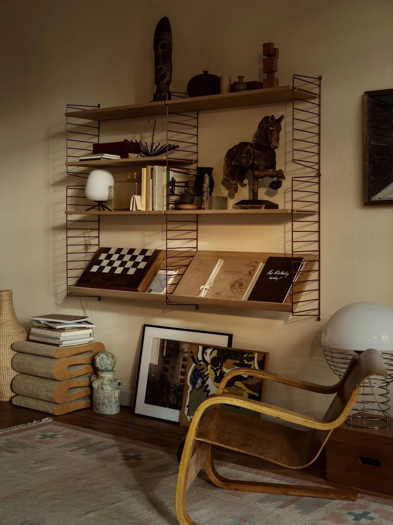 Ein gemütlicher Raum mit einem Regal, gefüllt mit Büchern, Kunst und Dekoration, neben einem Holz-Biegesessel und einer großen weißen Lampe.