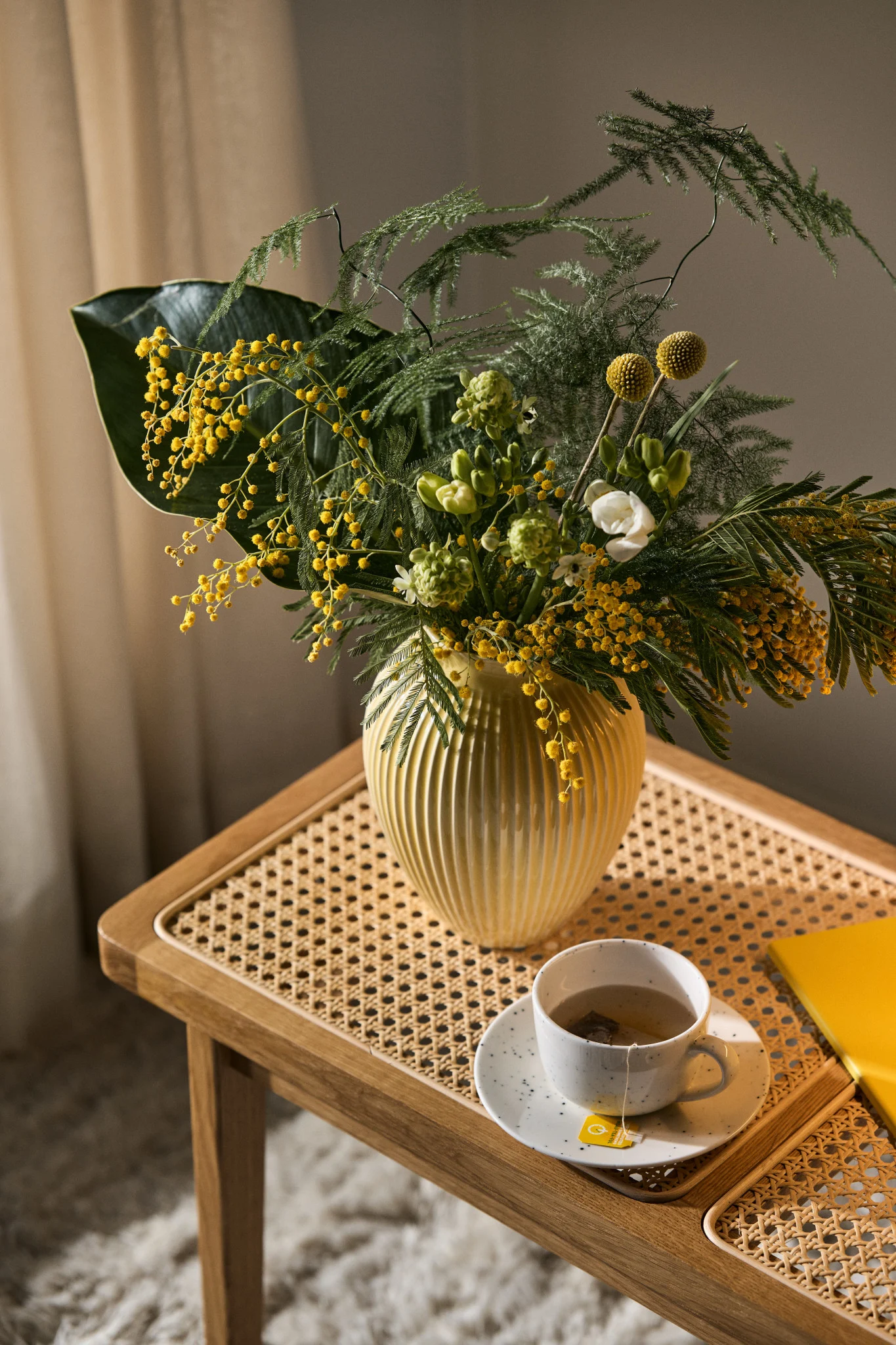 Auf einer Holzbank stehen eine Tasse Kaffee und ein hübsch arrangierter Blumenstrauß in einer geriffelten gelben Vase von Knabstrup.