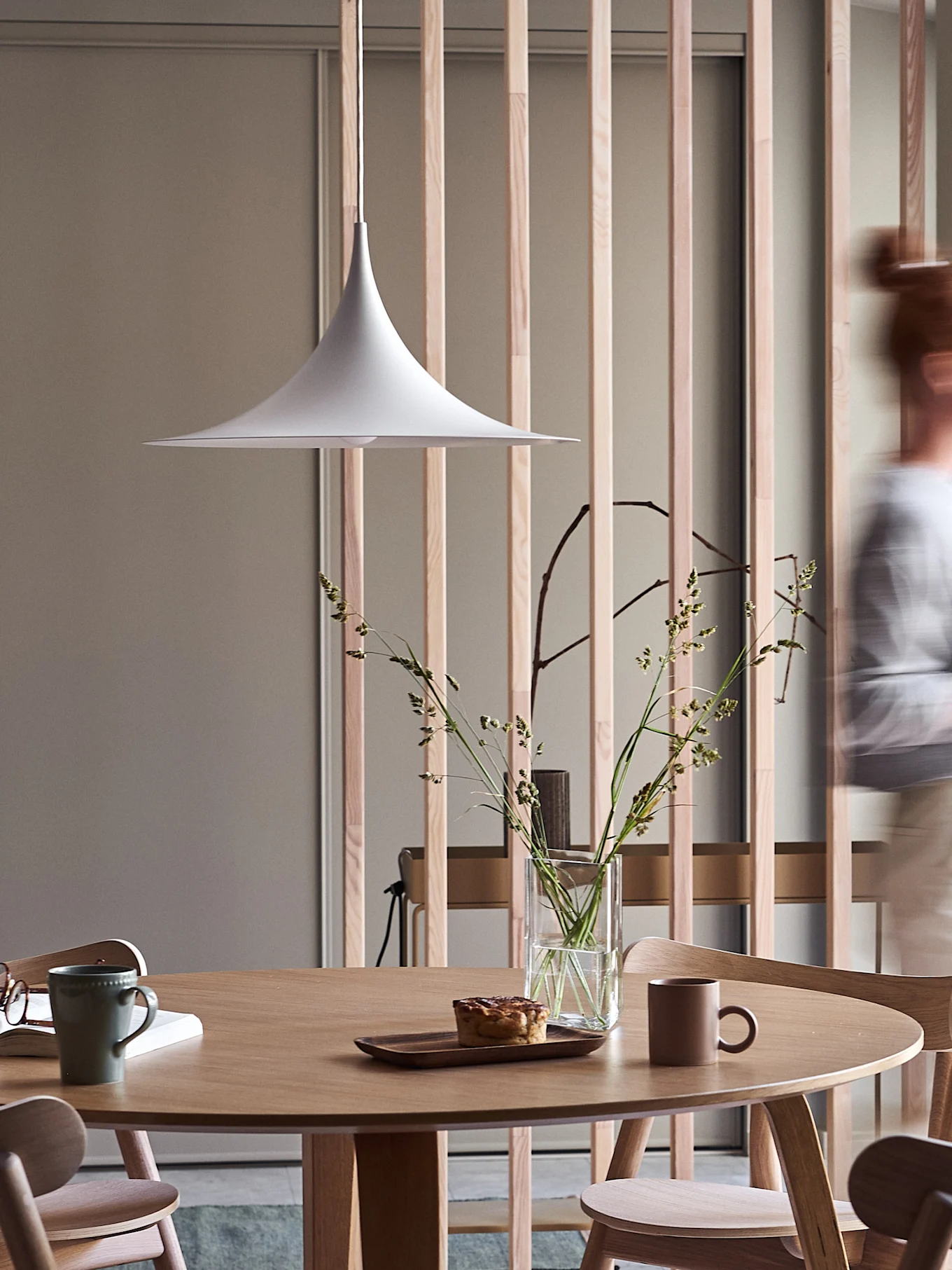 Moderner Essbereich mit rundem Holztisch, weißer Pendelleuchte, Vase mit Gräsern, Gebäck und Tassen, mit einer unscharfen Person im Vorbeigehen.