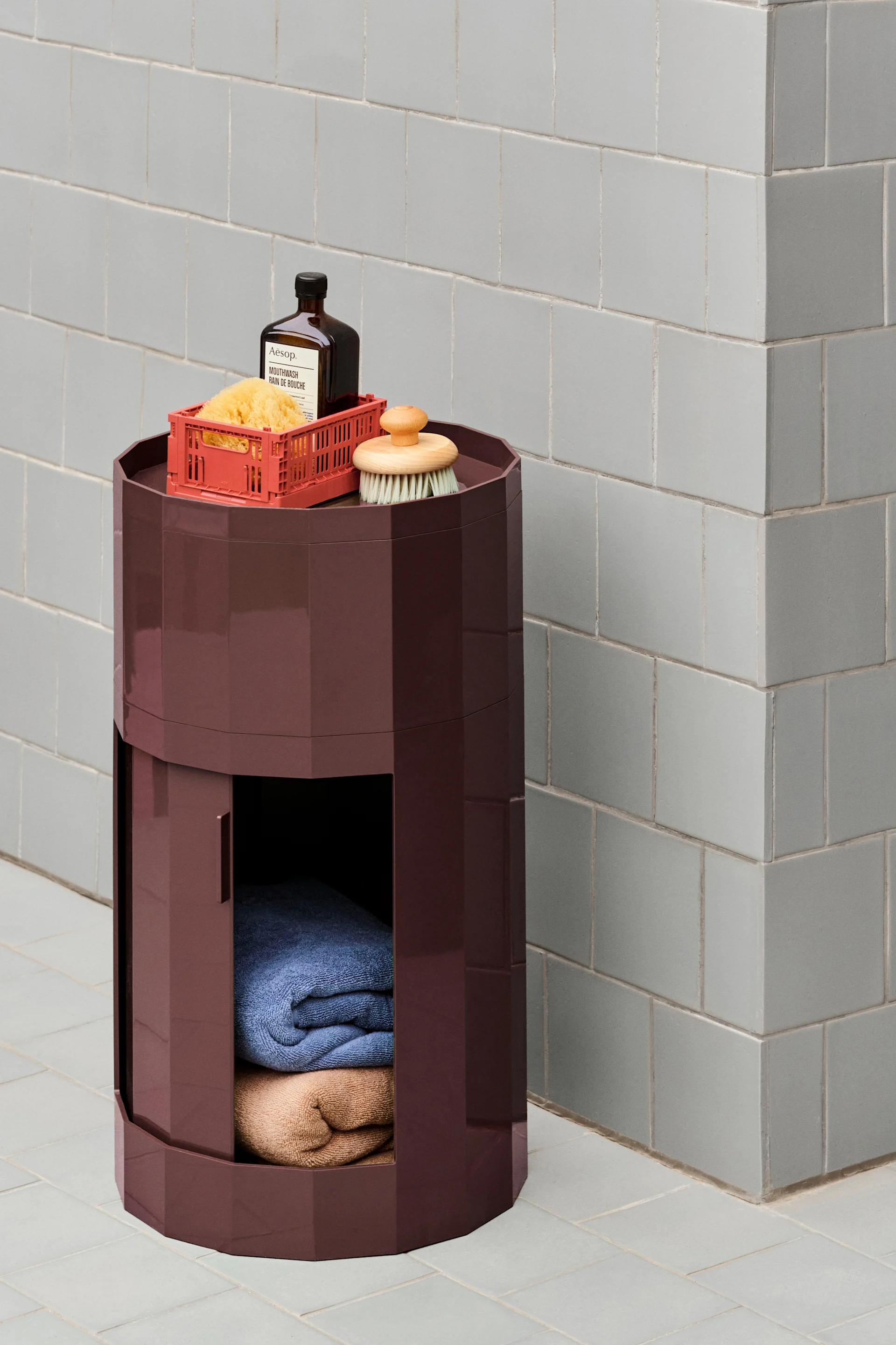 Ein dunkelrotes, sechseckiges Aufbewahrungsmöbel mit Badeprodukten oben und gefalteten Handtüchern innen, vor einer hellgrau gefliesten Wand.