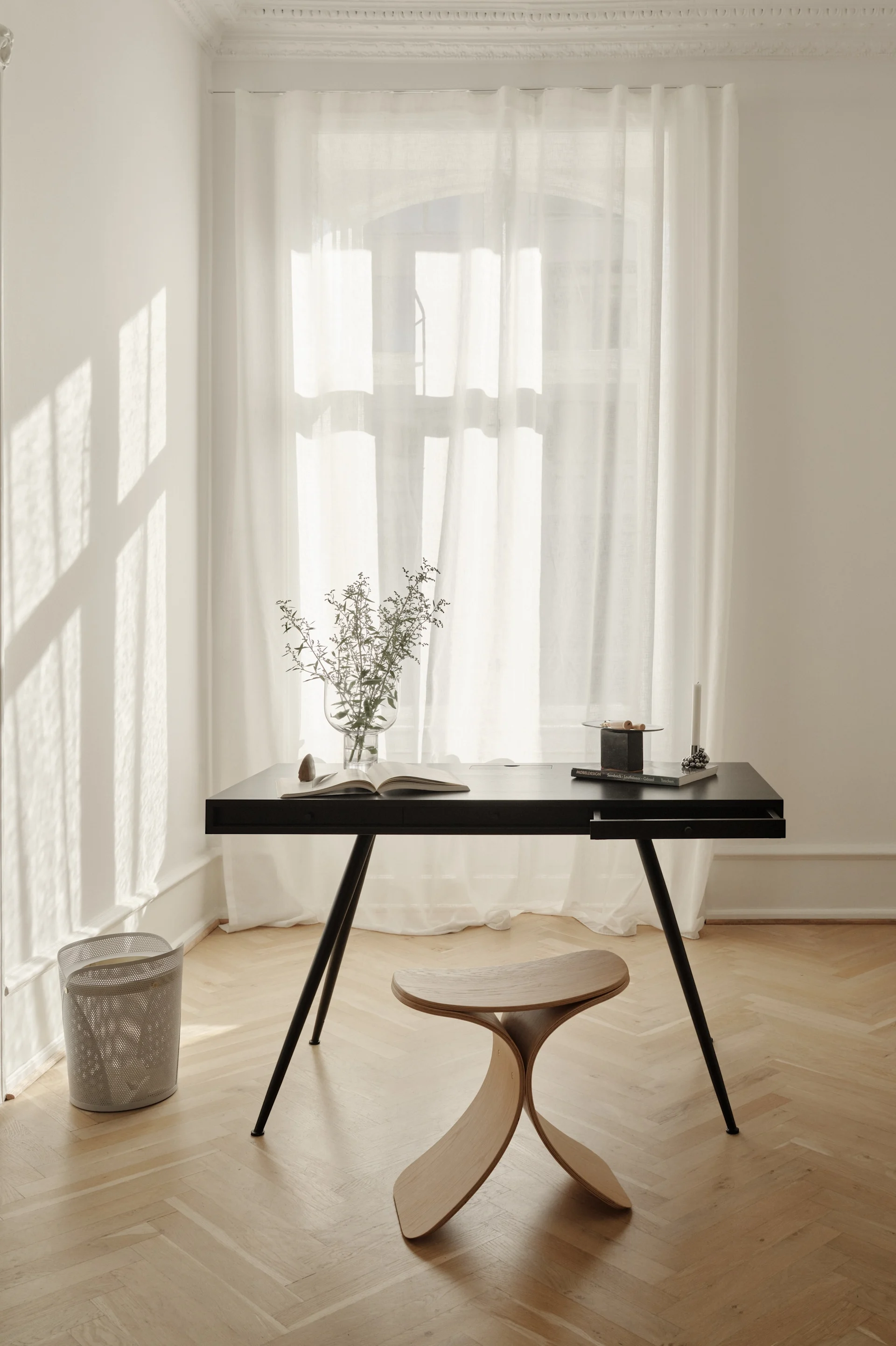 Ein minimalistisches Heimbüro mit einem schwarzen Schreibtisch, einem geschwungenen Holzhocker, einer Vase mit Blattwerk und einem offenen Buch an einem Fenster mit transparenten Vorhängen.