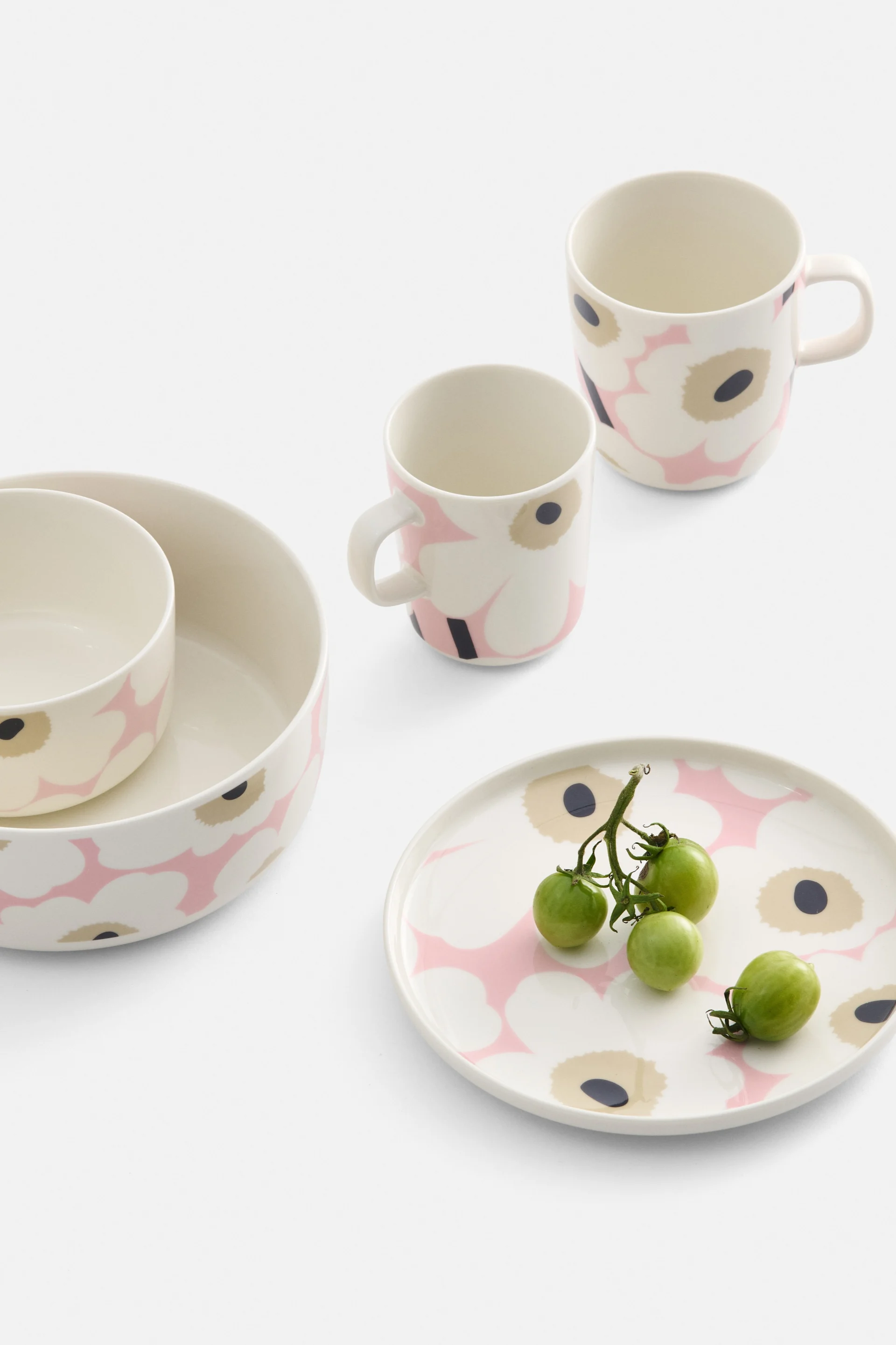 Cremefarbenes Geschirr mit rosa und schwarzem Blumenmuster, darunter zwei Schalen, zwei Tassen und ein Teller mit grünen Kirschtomaten.