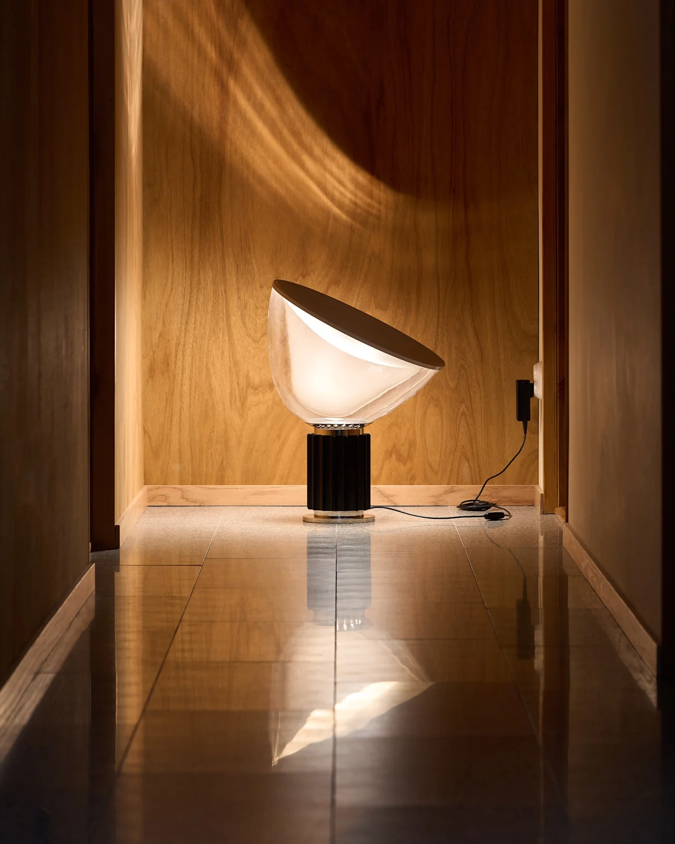 Moderne Lampe mit halbrunder, weißer Schirm und schwarzem, geriffeltem Sockel in Holzflur mit reflektierendem Boden. Lichtmuster an der Wand.