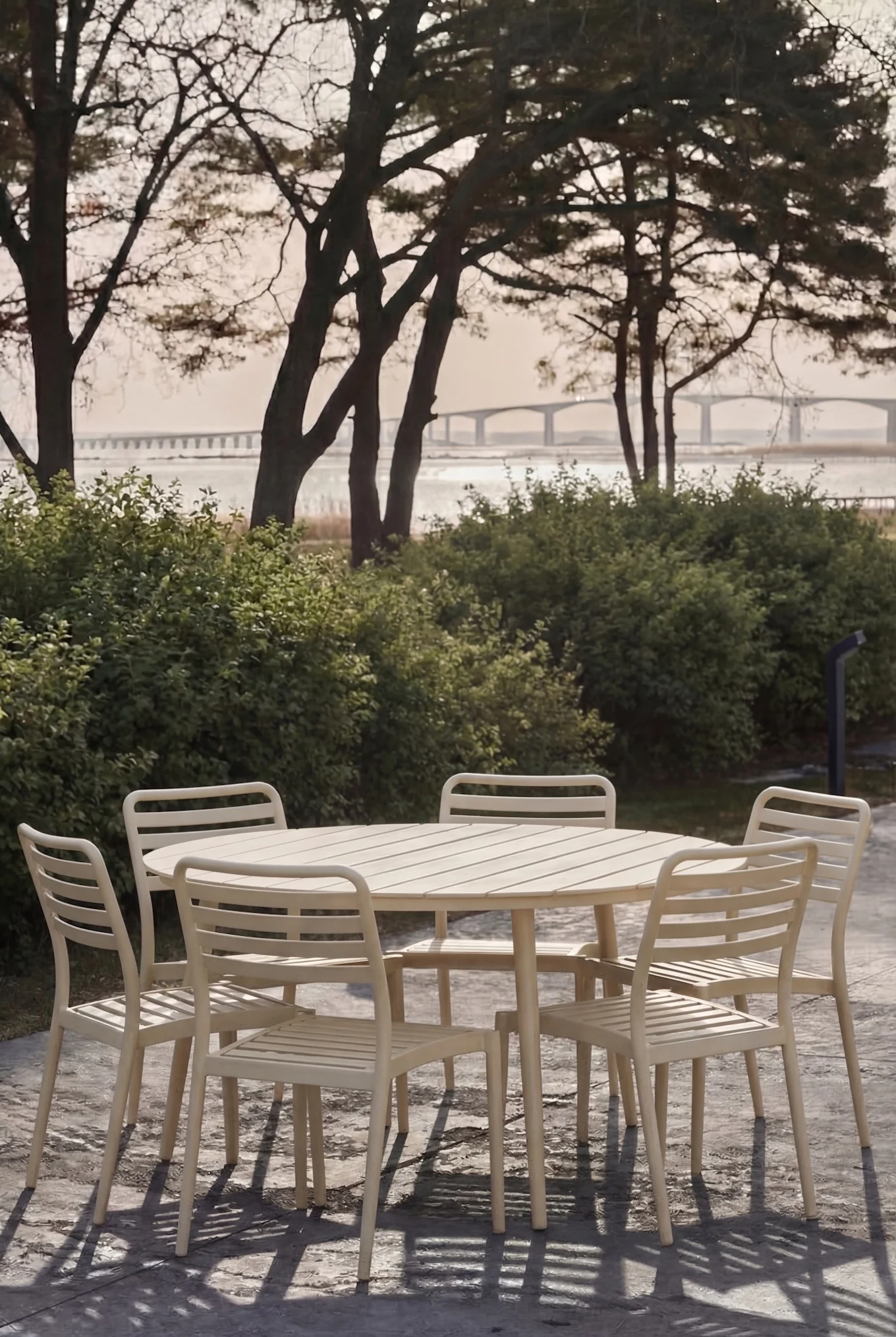Helles Gartenset mit rundem Lamellentisch und sechs Stühlen auf Terrasse, Bäume und Brücke im Hintergrund.