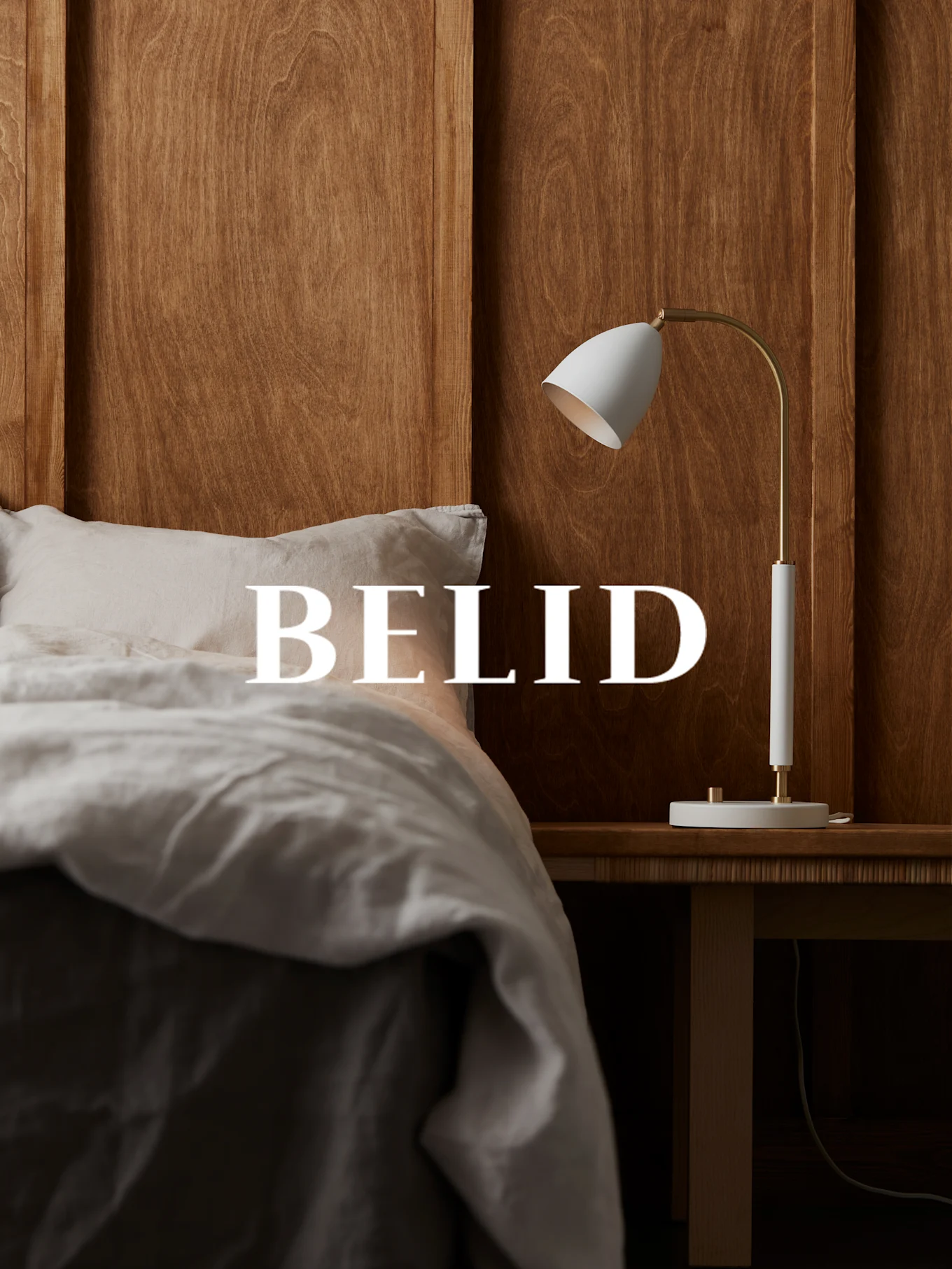 Weiße und messingfarbene Lampe auf einem Nachttisch aus Holz, mit dem Text 'BELID' über einem Bett mit Leinenbettwäsche.