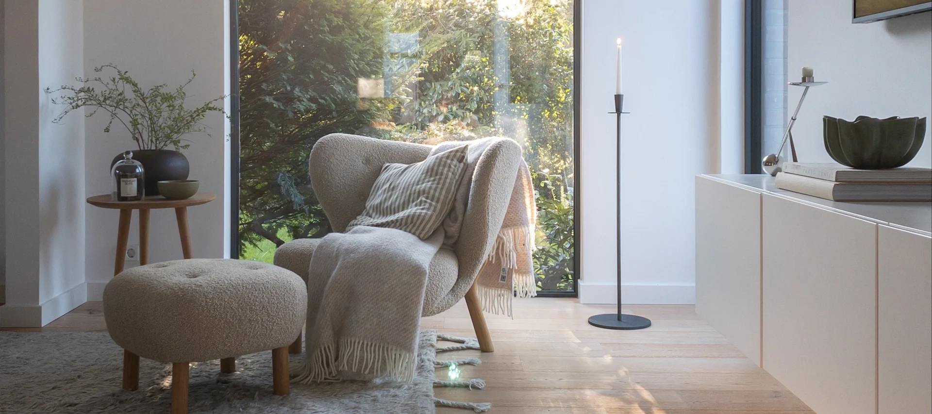 Gemütliche Wohnzimmerecke mit hellem Sessel, Fußhocker, Beistelltisch mit Deko, hohem Kerzenständer an Fenster zum Garten.